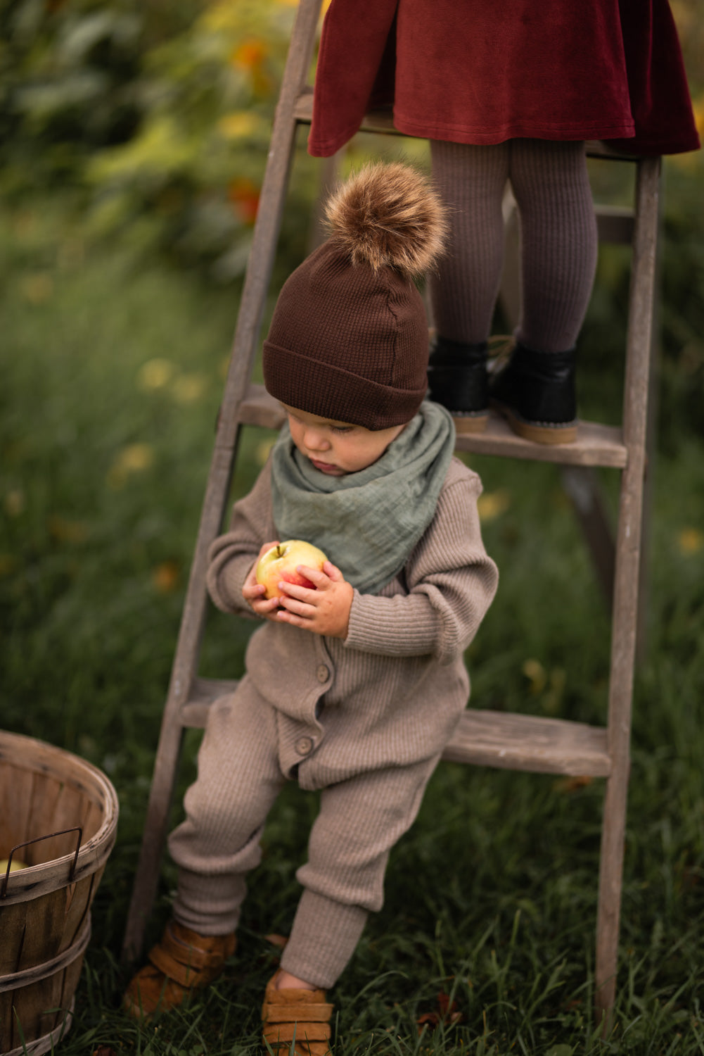 Mini Overall Rippstrick - Hafer: Ein Kind steht auf einer Leiter und hält einen Apfel, trägt den weichen, gerippten Overall aus recycelter Baumwolle, ideal für ganzjährigen Komfort und Anpassung.