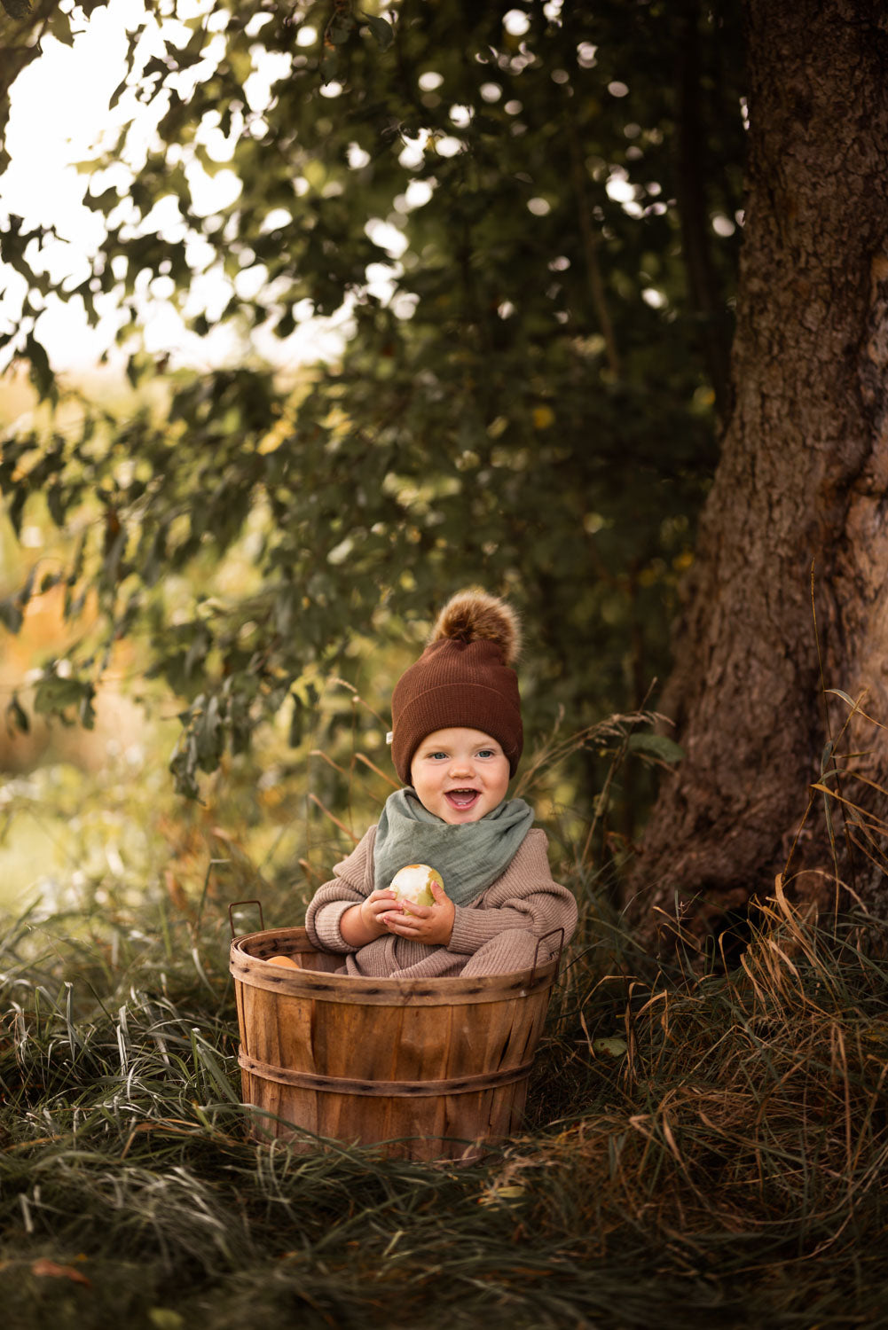 Baby in einem Korb hält einen Apfel, trägt den Mini Overall Rippstrick - Hafer, ideal für ganzjährigen Komfort und Nachhaltigkeit aus recycelter Baumwolle. Der perfekte Strampler für Babys.