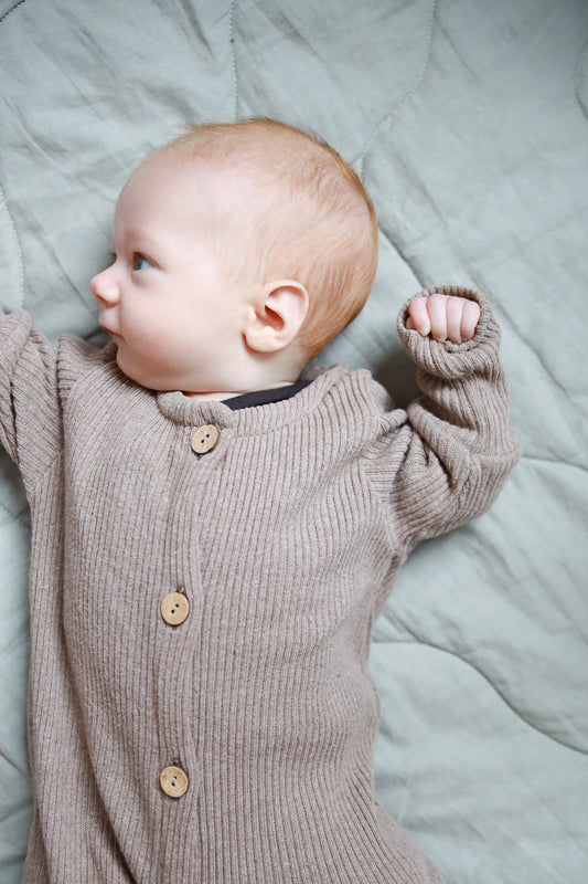 Baby liegt auf einer Decke in einem Mini Overall Rippstrick - Hafer. Der Overall aus recycelter Baumwolle bietet Komfort und wächst dank Krempelbündchen mit. Perfekt für alle Jahreszeiten.  Erstausstattung Baby und Baby Geschenk zur Geburt.
