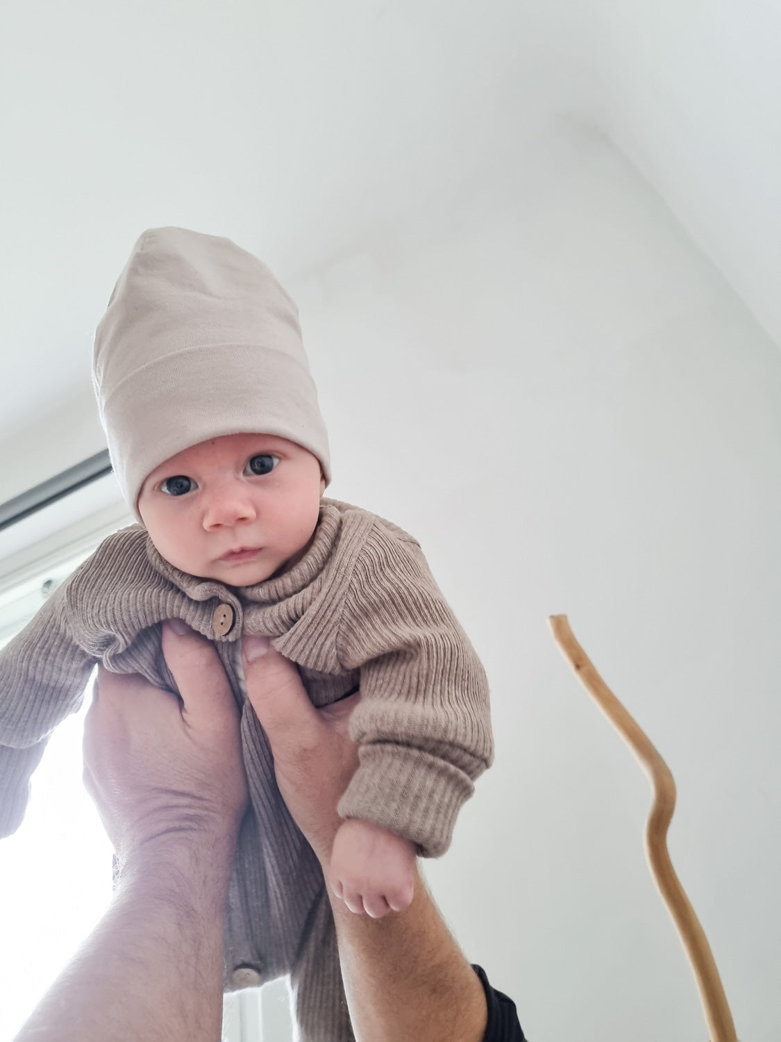 Baby trägt Mini Overall Rippstrick - Hafer. Ein Strampler für Babys aus recycelter Baumwolle mit Beanie aus Kaschmir Mix, ideal für ganzjährigen Komfort und Wachstum.