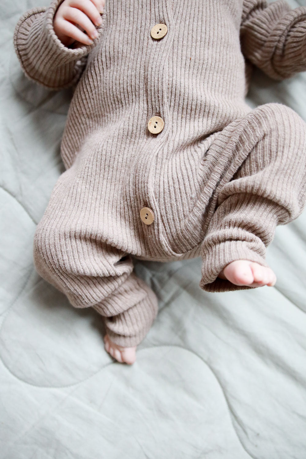 Baby in einem braunen Mini Overall Rippstrick - Hafer, aus recycelter Baumwolle mit Knöpfen auf gestricktem Stoff, liegend auf einem Bett. Der Overall bietet Komfort und wächst dank Krempelbündchen mit.  Erstausstattung Baby und Baby Geschenk zur Geburt.