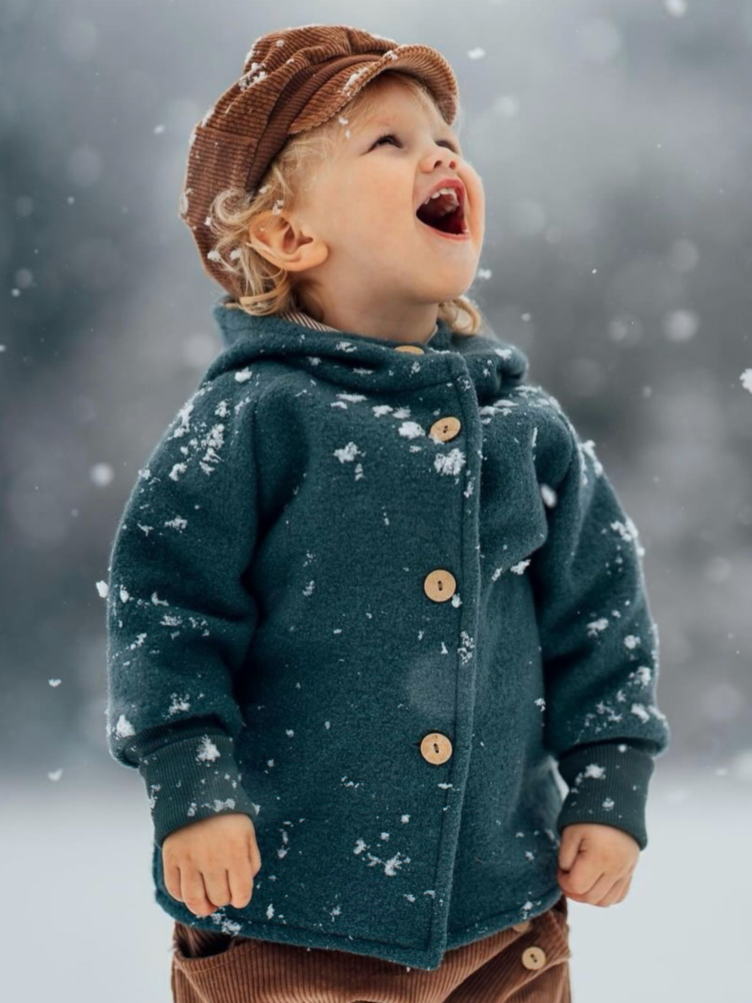 Ein Kind trägt die Michelmütze Breitcord Stretch - Weizen, während es im Schnee steht. Die Mütze mit Schirm und Zugband schützt vor Wind und wächst mit, ideal für aktive Abenteuer.