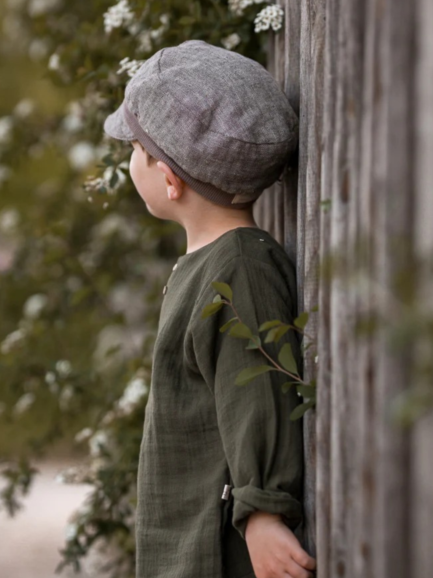 Kind mit Michelmütze Fischgrat Maus in Profilansicht an einem Holzzaun. Die Mütze hat ein Fischgrätmuster, einen kleinen Schirm und verstellbares Zugband, passend für Kinderköpfe.