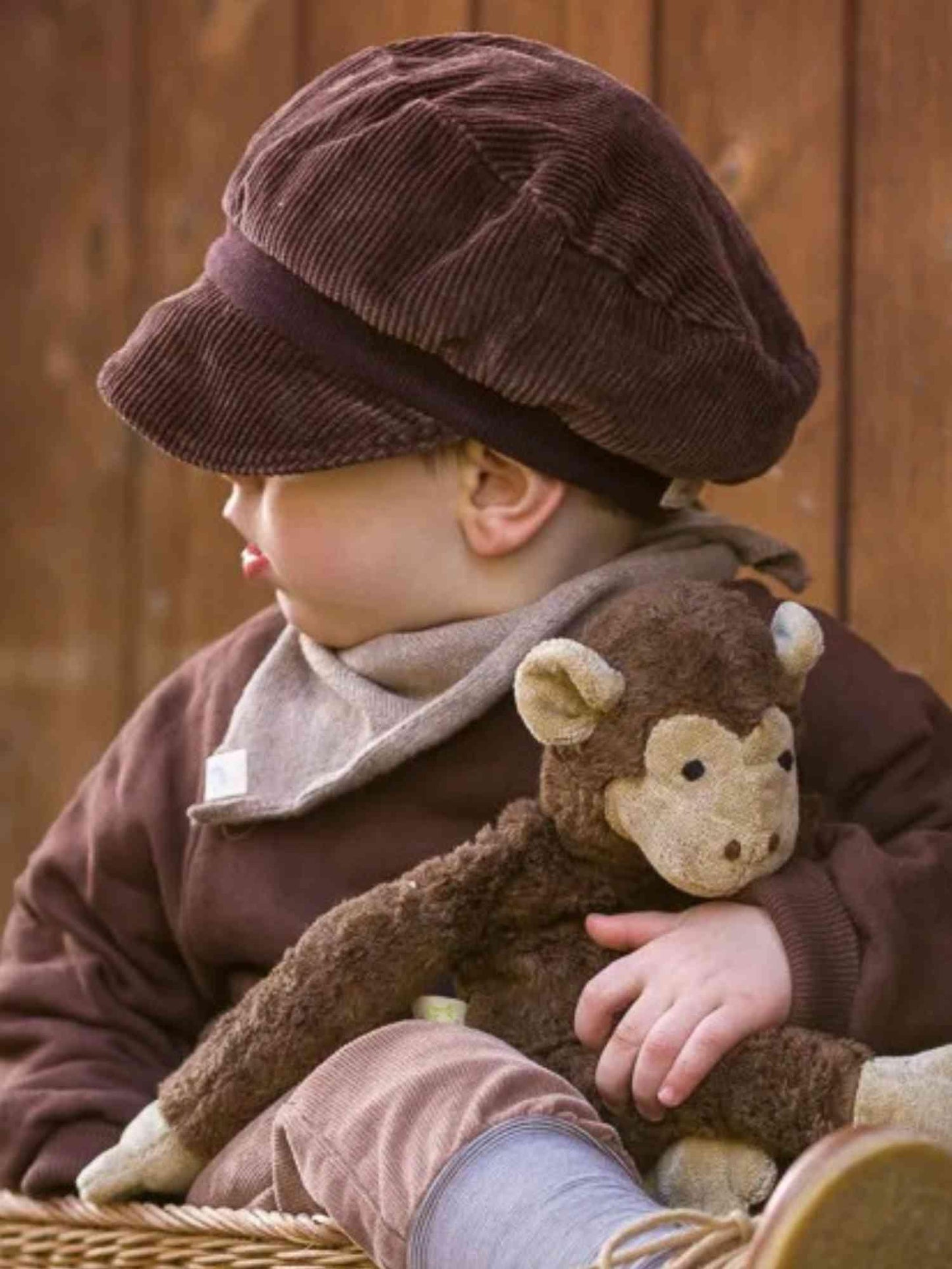 Kleinkind mit Michelmütze Breitcord - Schoko, einer weichen, elastischen Cord-Kappe mit Bio-Baumwollbündchen, sitzt und hält ein Plüschtier, die Mütze verdeckt teilweise die Augen.