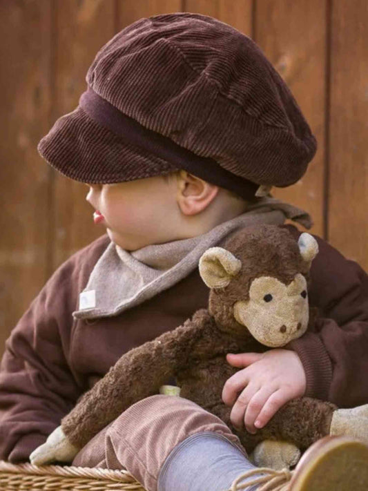 Kleinkind mit Michelmütze Breitcord - Schoko, einer weichen, elastischen Cord-Kappe mit Bio-Baumwollbündchen, sitzt und hält ein Plüschtier, die Mütze verdeckt teilweise die Augen.