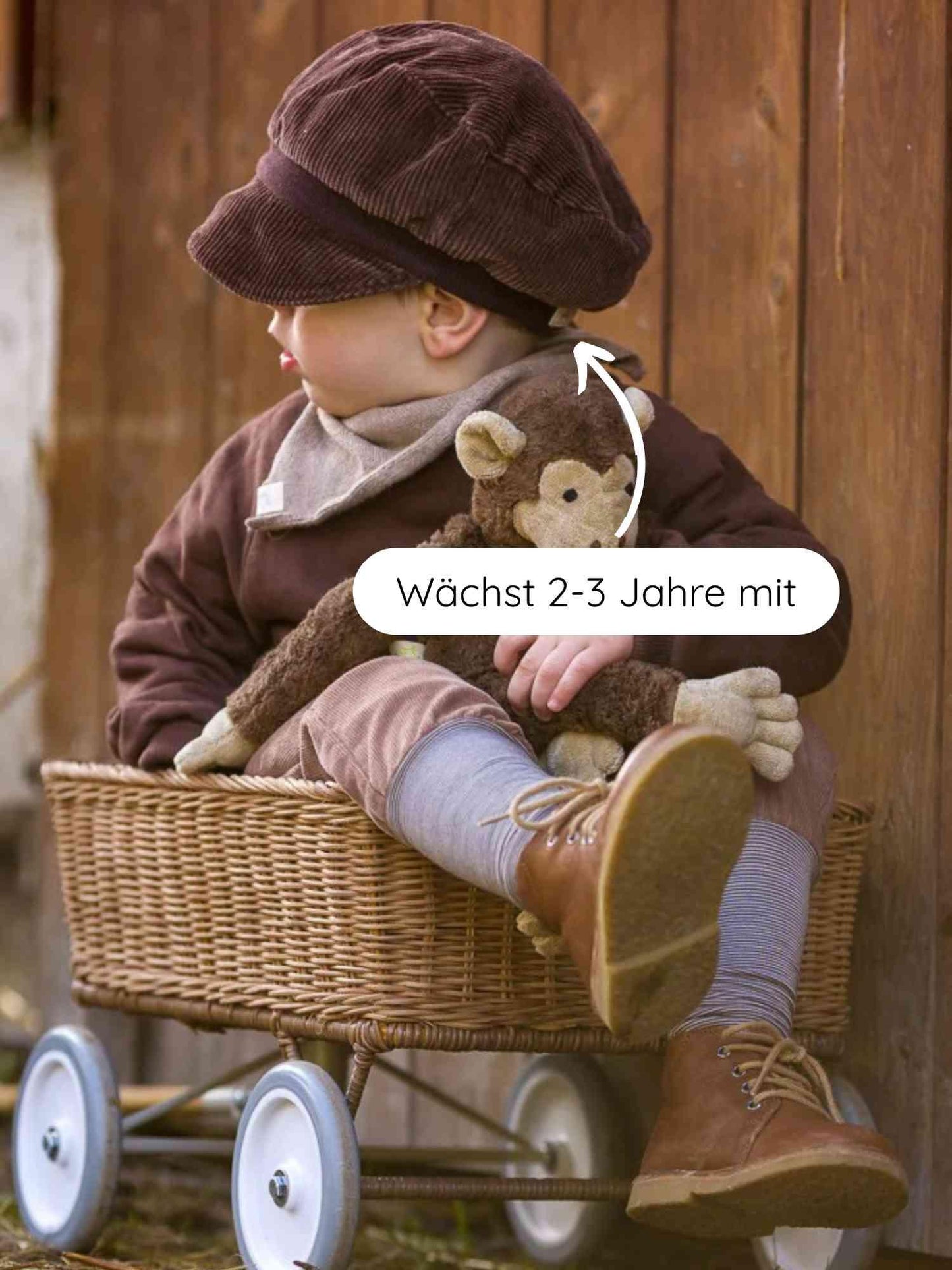 Ein Kleinkind sitzt im geflochtenen Wagen, trägt die Michelmütze Breitcord - Schoko. Die Mütze wächst 2-3 Jahre mit, ist aus robustem Cord mit Bio-Baumwollbündchen und sitzt bequem.