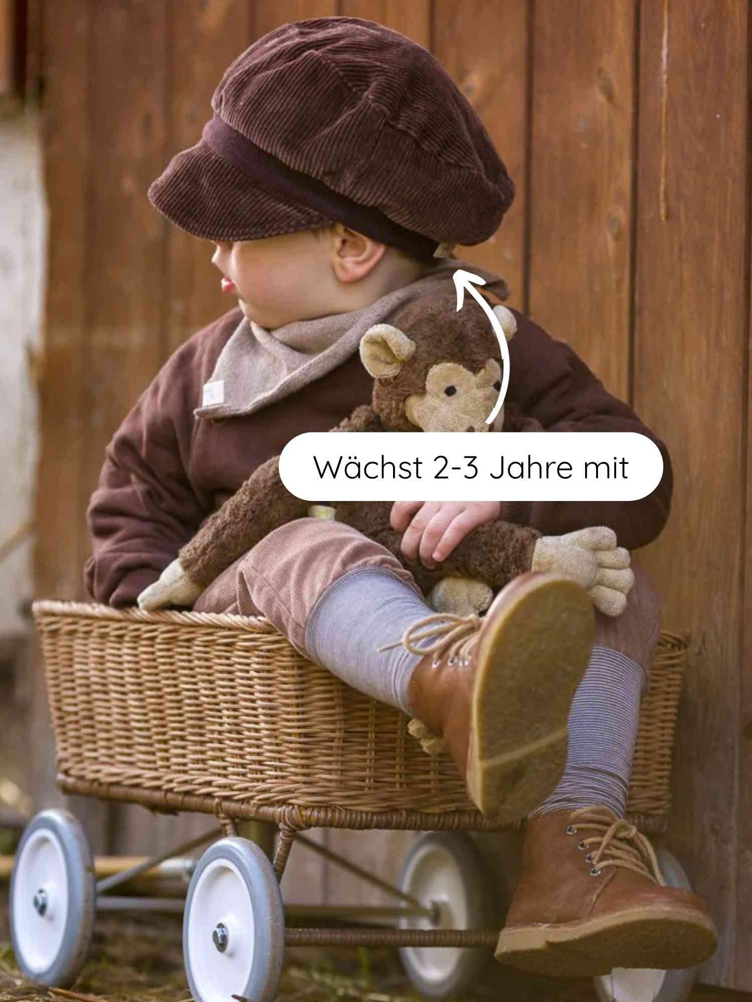 Ein Kleinkind sitzt im geflochtenen Wagen, trägt die Michelmütze Breitcord - Schoko. Die Mütze wächst 2-3 Jahre mit, ist aus robustem Cord mit Bio-Baumwollbündchen und sitzt bequem.