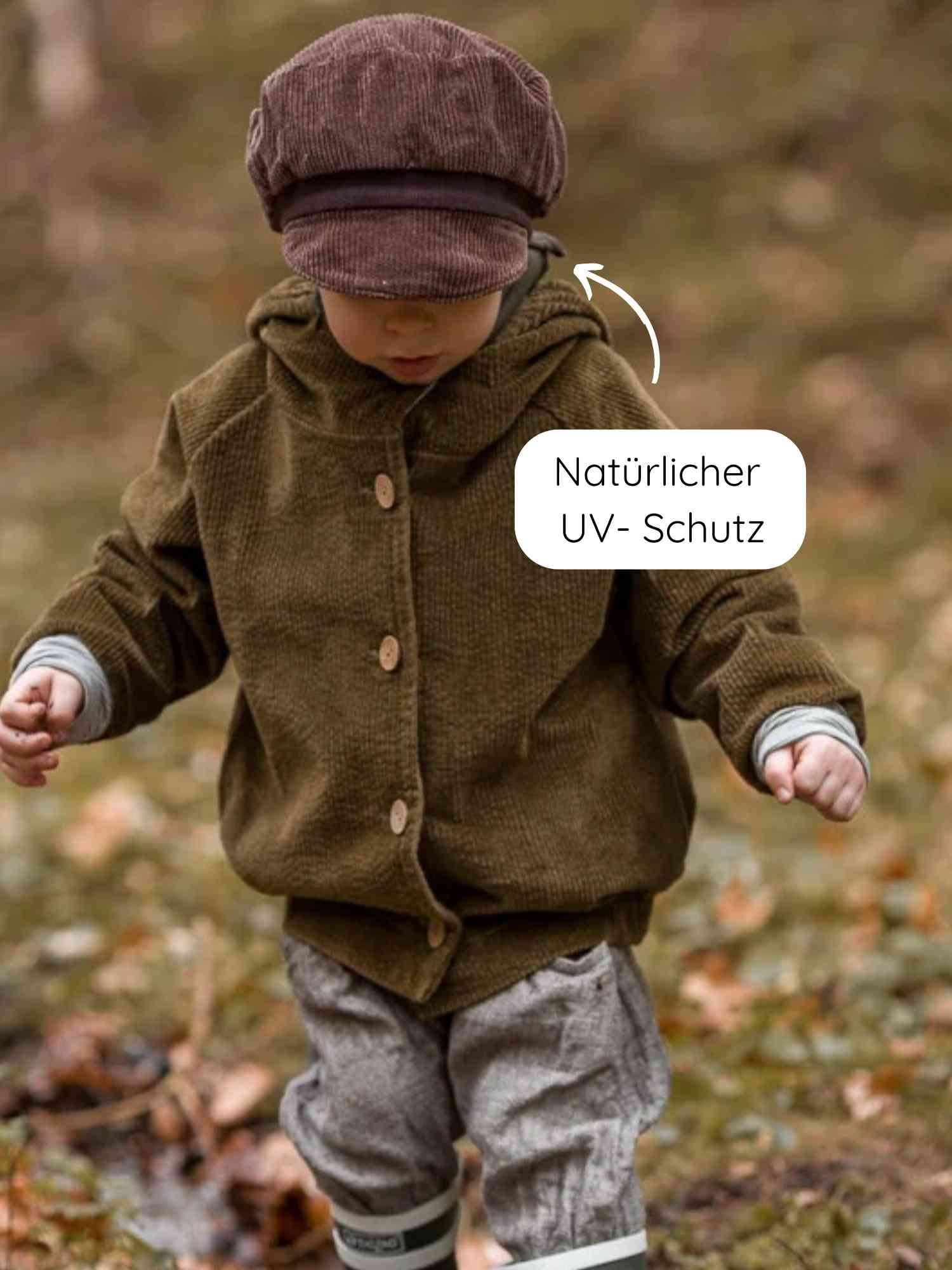 Ein Kleinkind trägt die Michelmütze Breitcord - Schoko, eine schicke braune Cordmütze mit verstellbarem Riemen, kombiniert mit einer Jacke und gestreiften Gummistiefeln auf einem herbstlichen Waldboden.