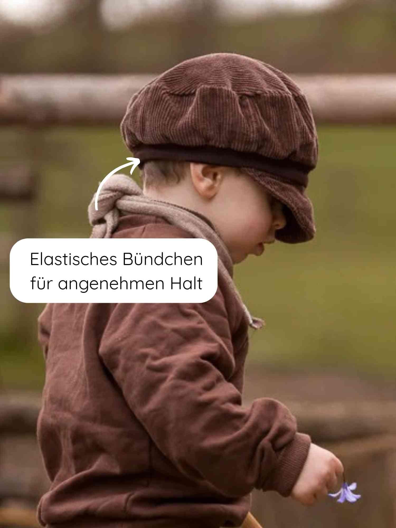Kind im Profil mit Michelmütze Breitcord - Schoko, zeigt elastisches Bündchen für angenehmen Halt. Hält lila Blume, trägt passende erdige Kleidung, steht an einem Holzzaun. Perfekt für ganzjähriges Tragen.