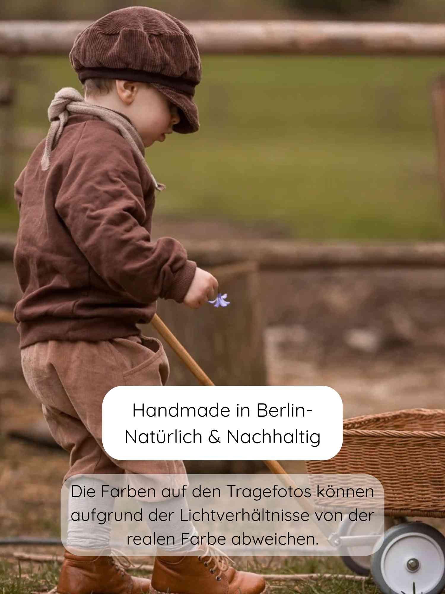 Ein Kleinkind trägt die Michelmütze Breitcord - Schoko, kombiniert mit einem erdtönigen Outfit. Es hält eine Blume und einen Holzgriff eines niedrigen Korbwagens, symbolisiert Nachhaltigkeit und Handarbeit.