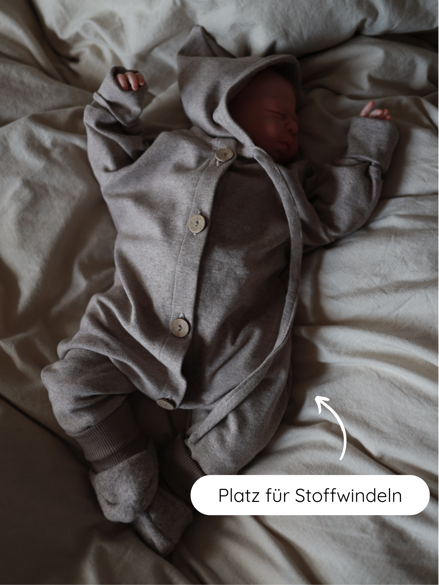 Neugeborenes im Mini Overall Feinstrick - Hafer Meliert, auf einer Decke liegend. Der Overall hat eine Kapuze und integrierte Booties, mit extra Platz für Stoffwindeln im Schrittbereich.