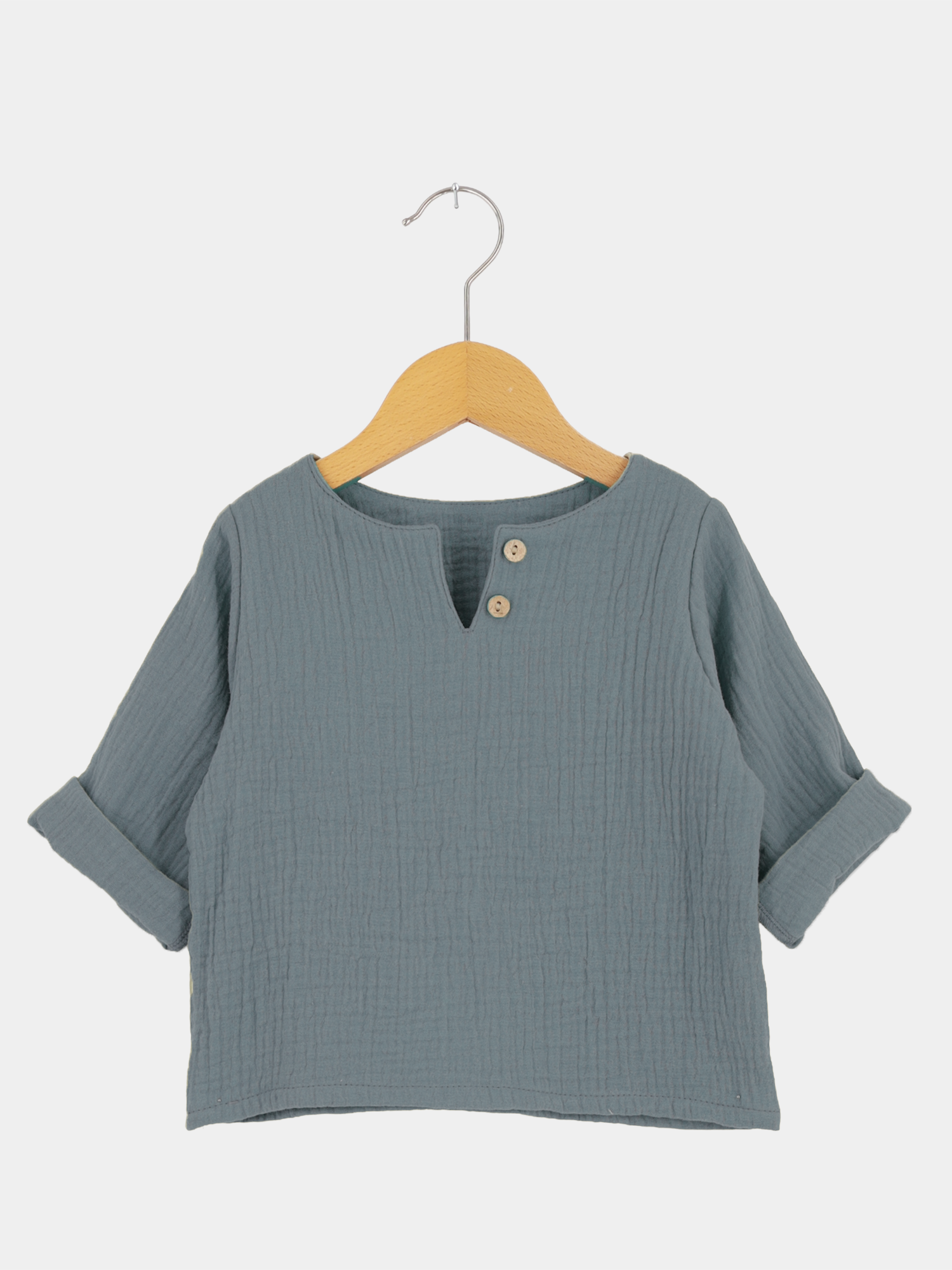 Hemdchen Musselin - Himmelblau: Langärmeliges Kinderhemd aus weichem, geknittertem Musselin mit Rundhalsausschnitt und Holzknöpfen, an einem Holzkleiderbügel hängend. Perfekt für warme Tage.