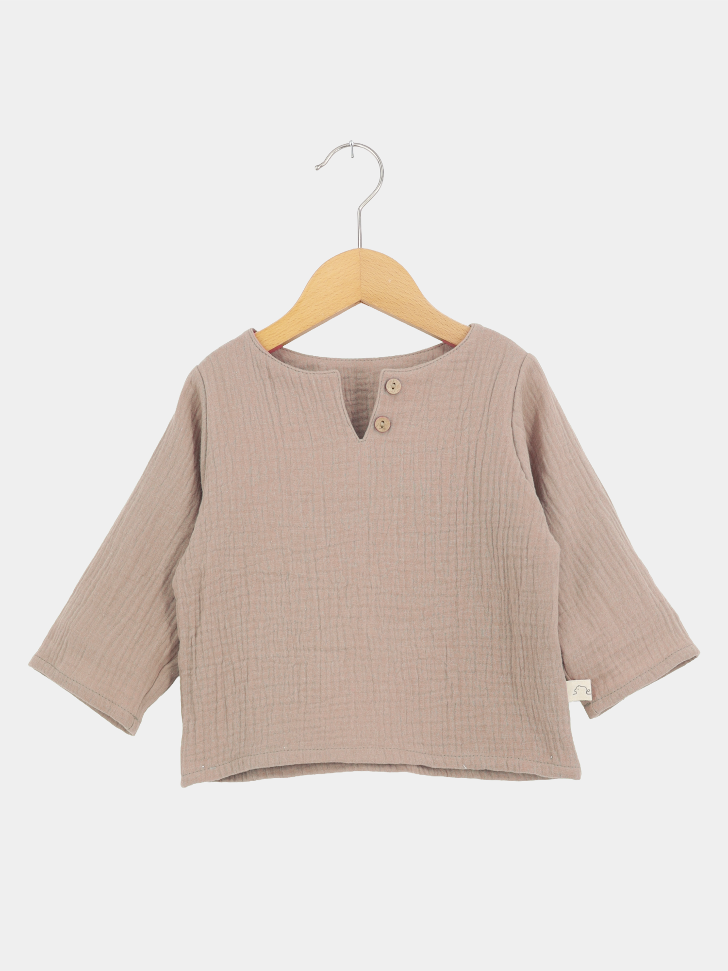 Hemdchen Musselin - Milchkaffee: Langarmshirt aus weichem, geknittertem Baumwollstoff mit V-Ausschnitt und Holzknöpfen, auf einem Holzkleiderbügel hängend, ideal für Babys und Kleinkinder.