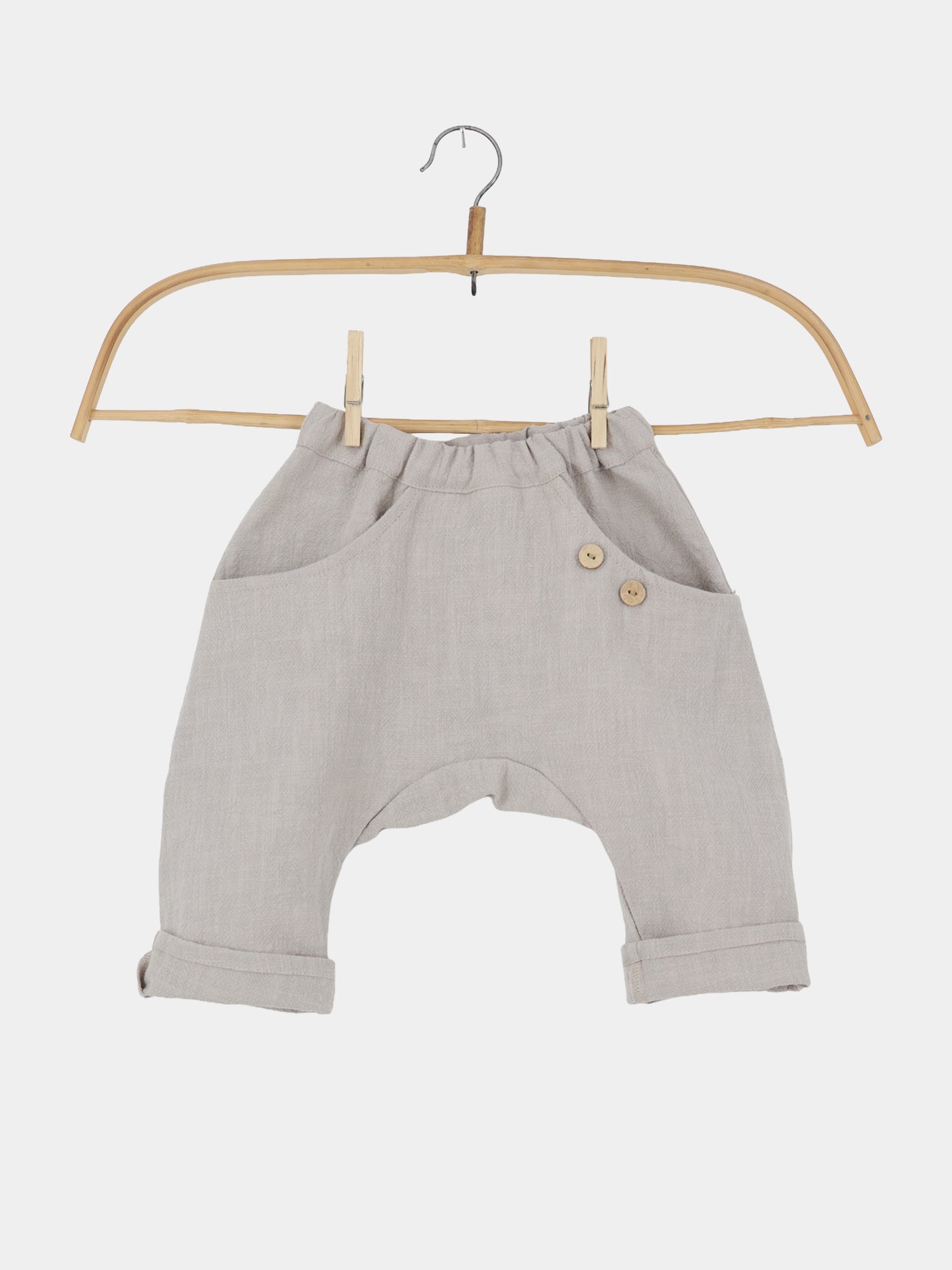 Sommerknicker Leinen - Sand: Leicht-beige, locker geschnittene Haremshose für Kleinkinder mit tiefem Schnitt, elastischem Bund, zwei Seitentaschen mit Holzknöpfen, an einem einfachen Holzbügel hängend. Perfekt für warme Sommertage.