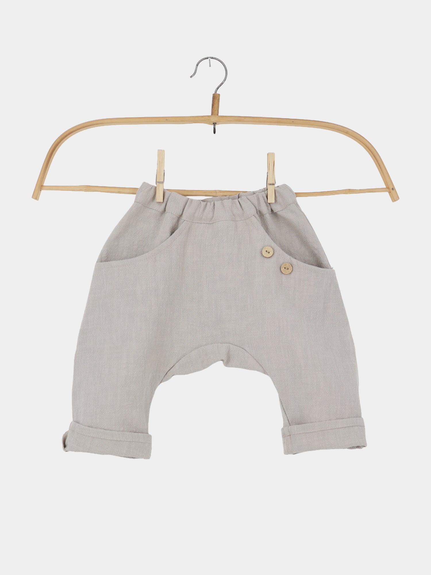 Sommerknicker Leinen - Sand: Leicht-beige, locker geschnittene Haremshose für Kleinkinder mit tiefem Schnitt, elastischem Bund, zwei Seitentaschen mit Holzknöpfen, an einem einfachen Holzbügel hängend. Perfekt für warme Sommertage.