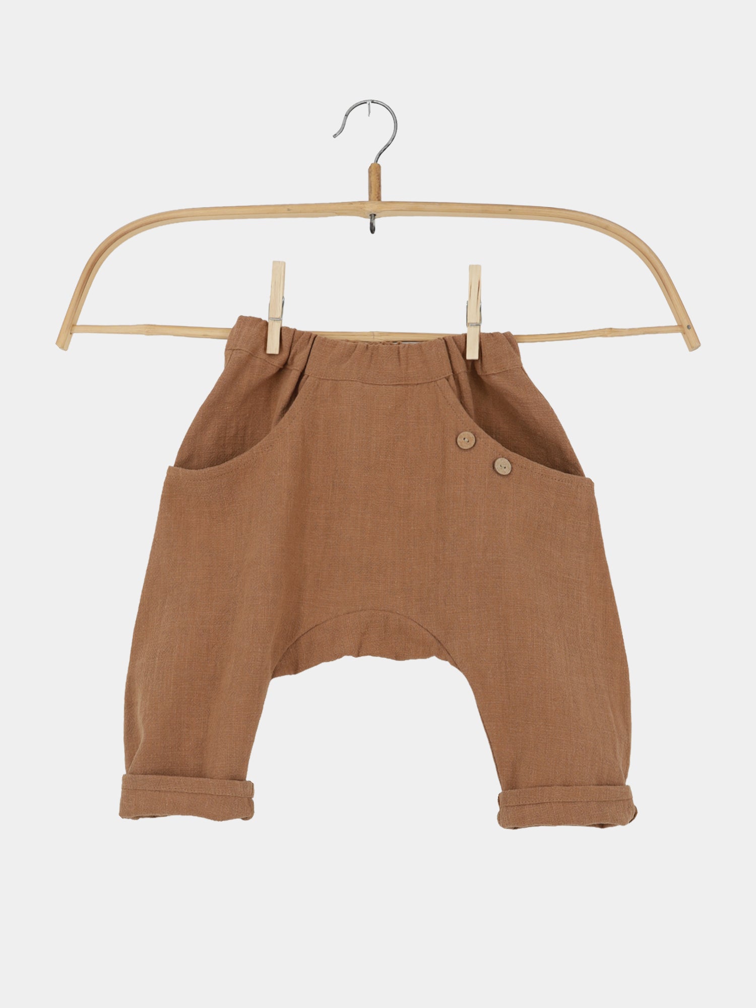 Sommerknicker Leinen - Weizen: Locker sitzende, braune Leinenhose für Kinder, mit elastischem Bund, zwei Fronttaschen, dekorativen Holzknöpfen und hochgerollten Beinabschlüssen, hängt an einem einfachen Holzkleiderbügel.