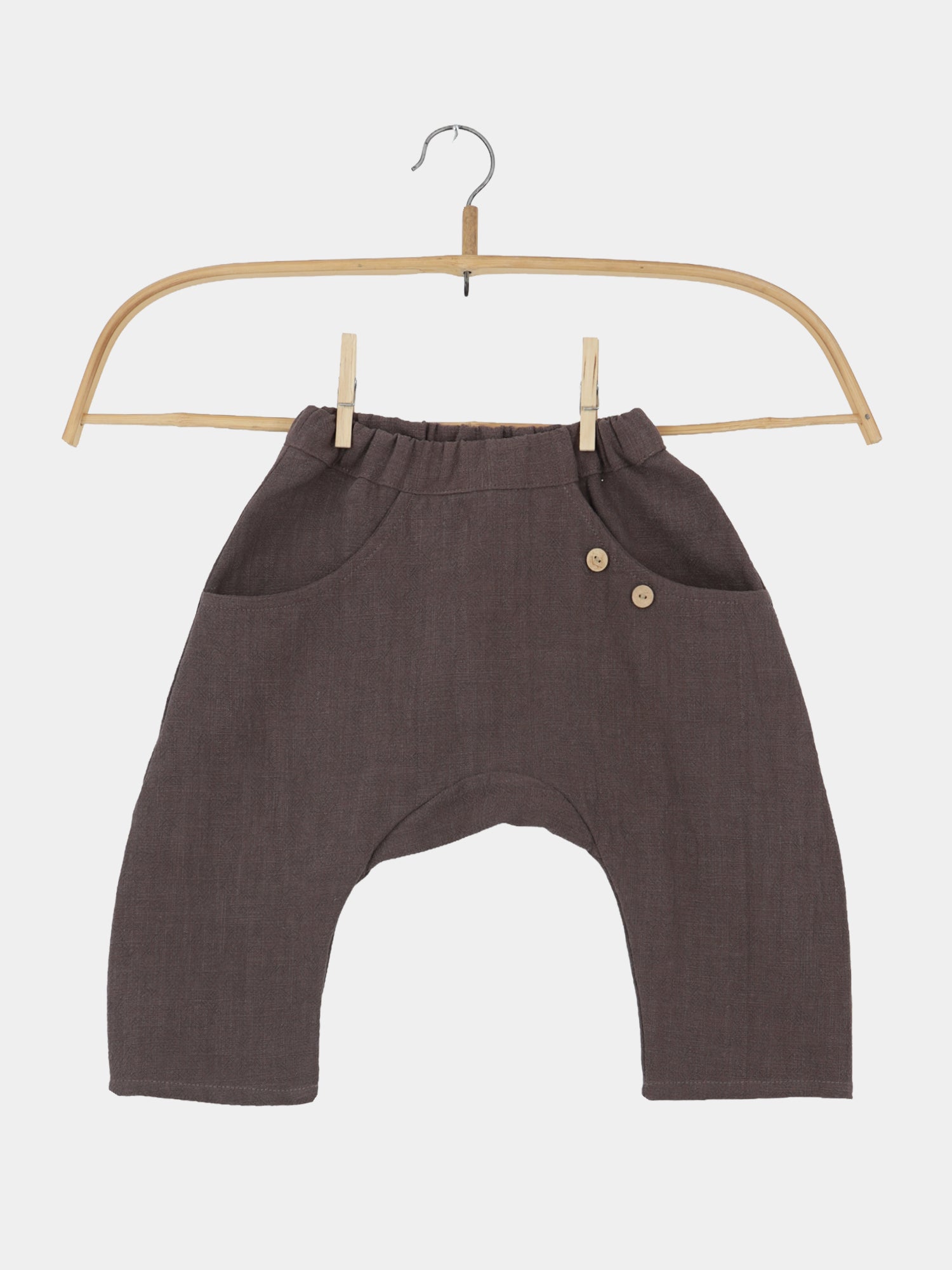 Sommerknicker Leinen - Erde: Locker geschnittene, taupefarbene Leinenhose mit elastischem Bund, seitlichen Taschen und dekorativen Holzknöpfen auf einem Holzkleiderbügel vor weißem Hintergrund. Ideal für aktive Kinder.