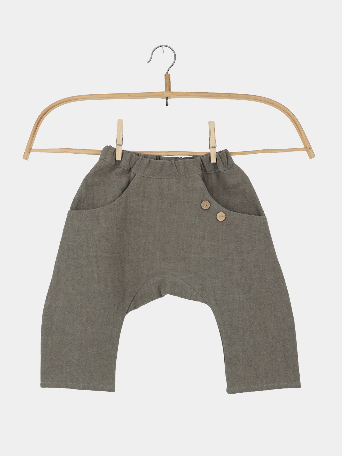 Sommerknicker Leinen - Oliv: Locker geschnittene, olivgrüne Kinderhose mit elastischem Bund, tiefem Schritt, zwei Fronttaschen mit Holzknöpfen, aus 100% Leinen, auf einem Holzkleiderbügel präsentiert.