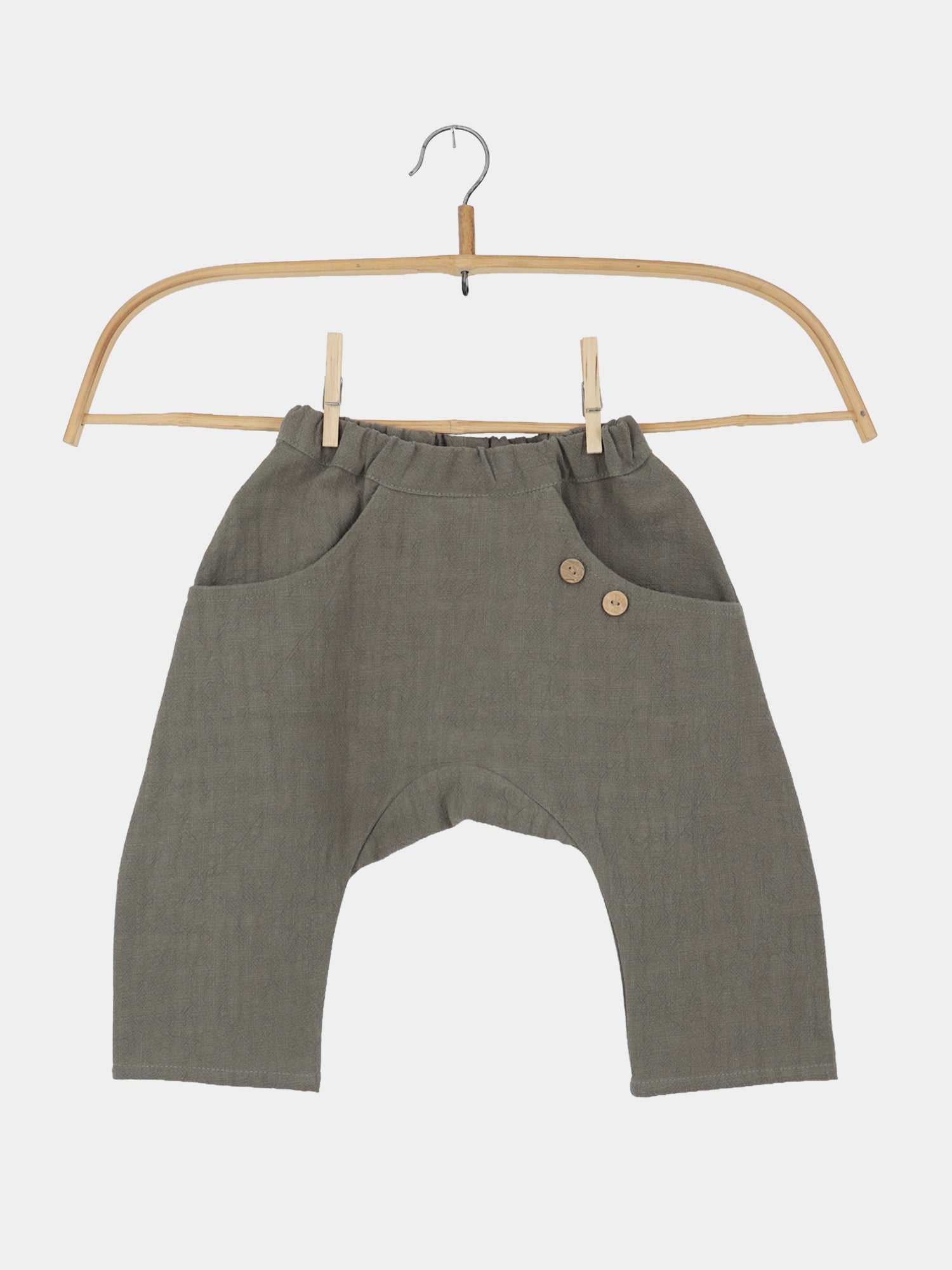 Sommerknicker Leinen - Oliv: Locker geschnittene, olivgrüne Kinderhose mit elastischem Bund, tiefem Schritt, zwei Fronttaschen mit Holzknöpfen, aus 100% Leinen, auf einem Holzkleiderbügel präsentiert.