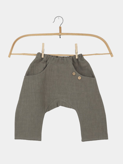Sommerknicker Leinen - Oliv: Locker geschnittene, olivgrüne Kinderhose mit elastischem Bund, tiefem Schritt, zwei Fronttaschen mit Holzknöpfen, aus 100% Leinen, auf einem Holzkleiderbügel präsentiert.