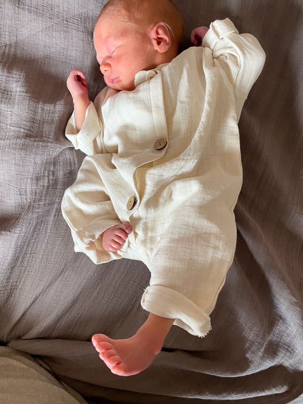 Ein Baby schläft in einem weißen Mini Overall Bio-Musselin, ideal für Sommer, auf einem Bett. Erstausstattung Baby