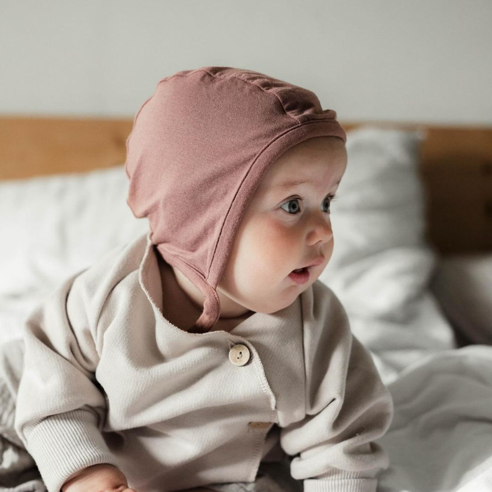 Baby sitzt auf dem Bett. Trägt Schlüttli und Baby Häubchen Kaschmir-Mix von Petit Cochon. Die perfekte Erstausstattung für Babys und Baby Geschenke.