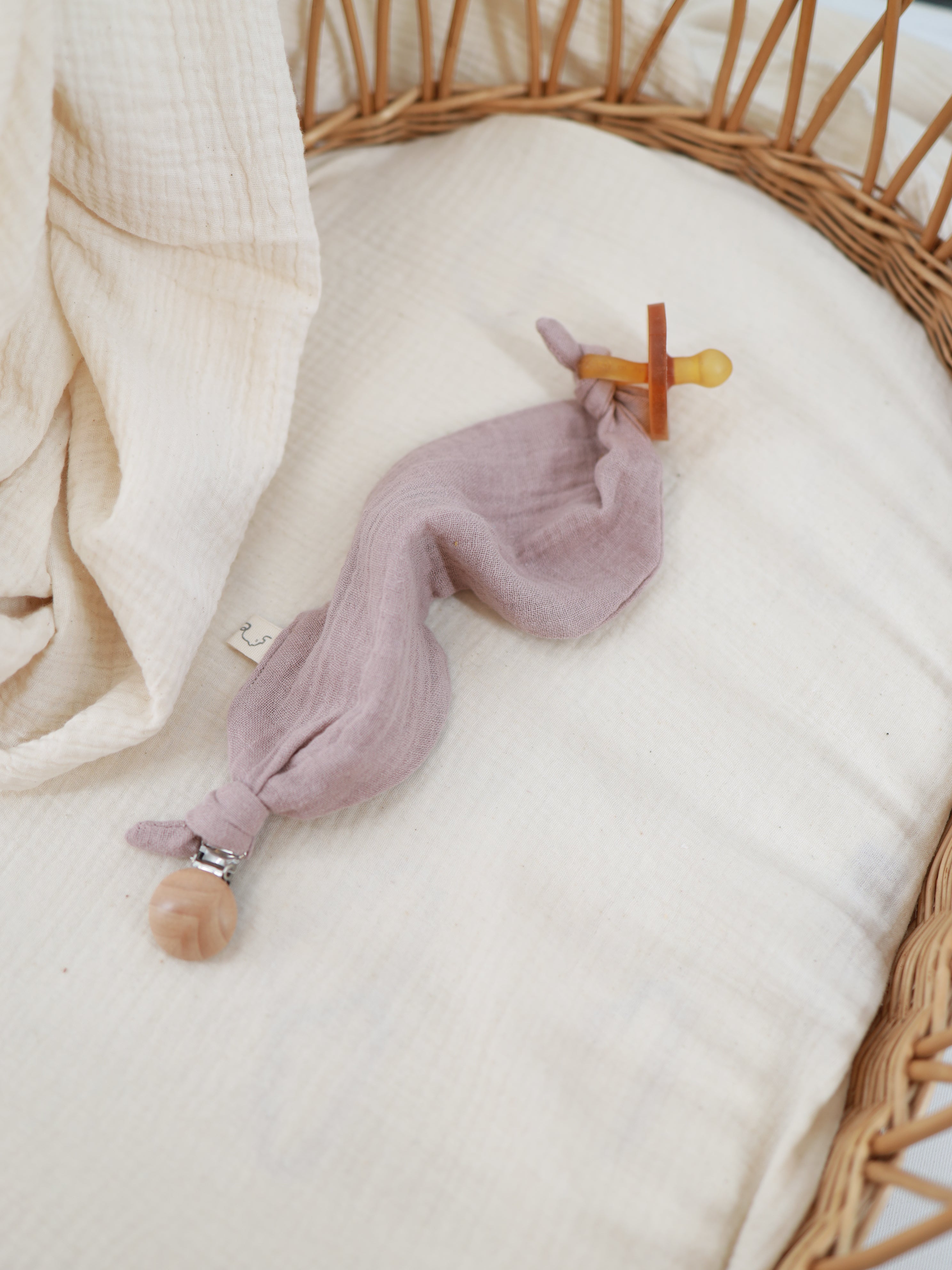Baby Schnuffeltuch /Schnullerband Baumwoll-Musselin - Puder: Ein Schnuller an einem Muslin-Schmusetuch, befestigt mit einem Holzperlenclip, auf einem cremigen Musselin-Liner in einem Korbbettchen.