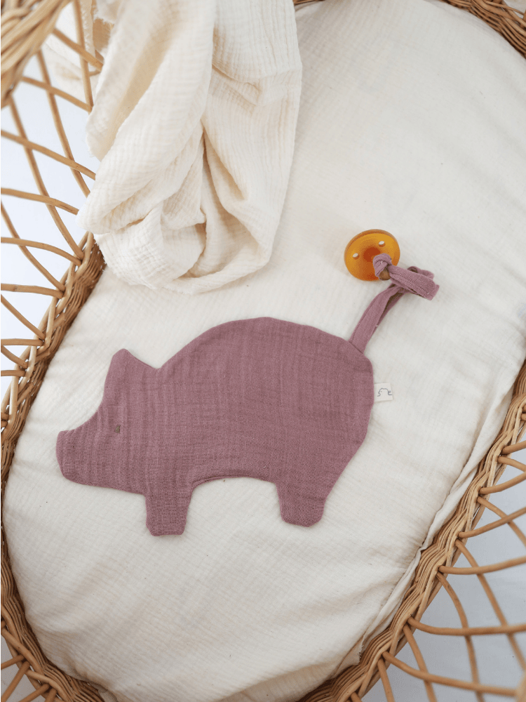 Schmusetuch/Schnullerband Schweinchen Baumwoll-Musselin - Malve in Babys Korb auf weichem Musselin, mit integriertem Schnullerband und liebenswertem Schweinchen-Design, ideal für sanfte Babypflege und Komfort.