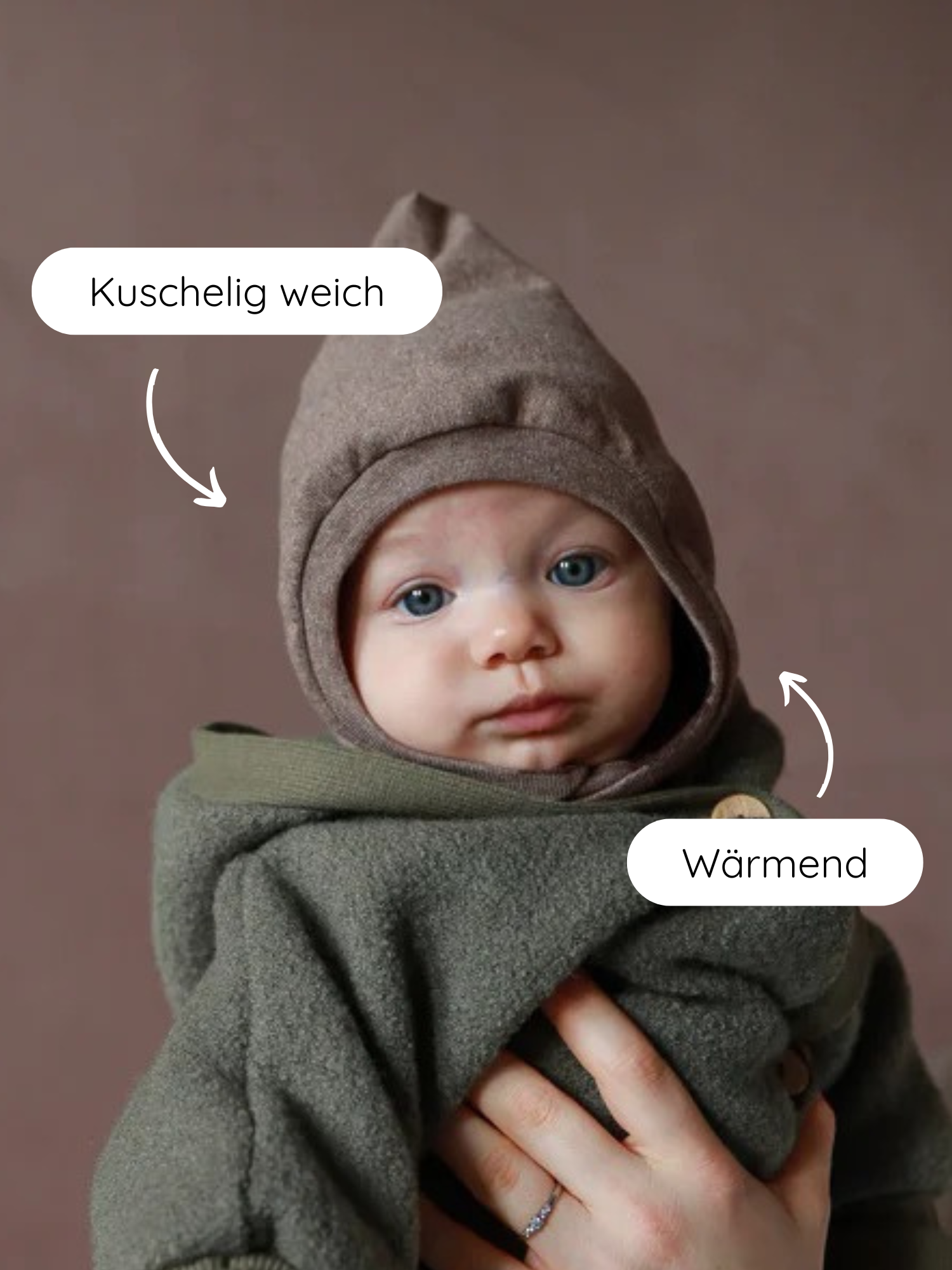 Ein Baby trägt die Schlupfmütze Feinstrick-Jersey - Praline Meliert, die in einem weichen, warmen, erdigen Ton gehalten ist. Ein Elternteil stützt es liebevoll, während es in einem olivegrünen Mantel gewärmt wird.