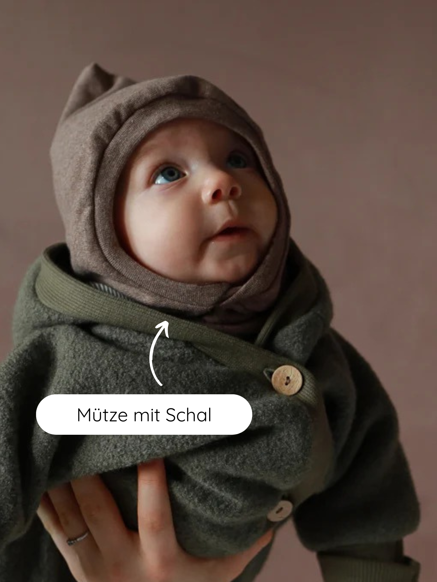 Ein Baby trägt die Schlupfmütze Feinstrick-Jersey - Praline Meliert, die den Kopf und Hals warm hält, kombiniert mit einer Wolljacke. Eine erwachsene Hand stützt das Baby sanft.