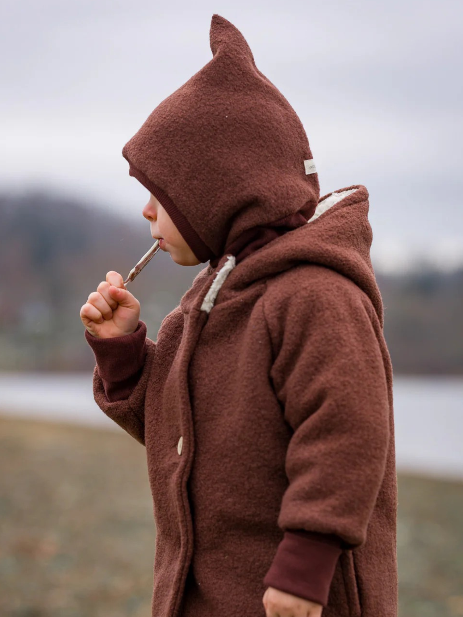 Ein Kind am See trägt die Schlupfmütze Wollwalk - Schoko, eine warme, atmungsaktive Kapuze aus Wollwalk, die Kopf und Hals an kühlen Tagen zuverlässig schützt.