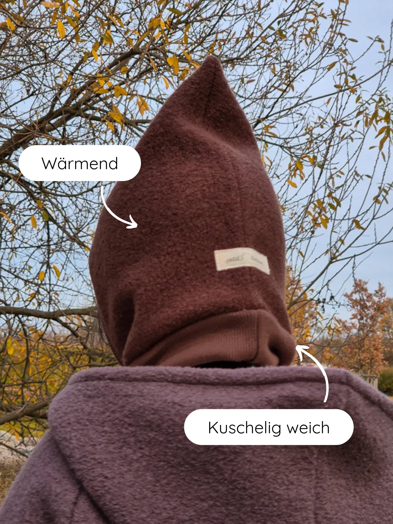 Person von hinten in einer Herbstlandschaft mit Schlupfmütze Wollwalk - Schoko, spitzem, braunem Hoodie, der wärmend wirkt, und kuschelig weichem, flauschigem Mantel in gedämpftem Pflaumenton.