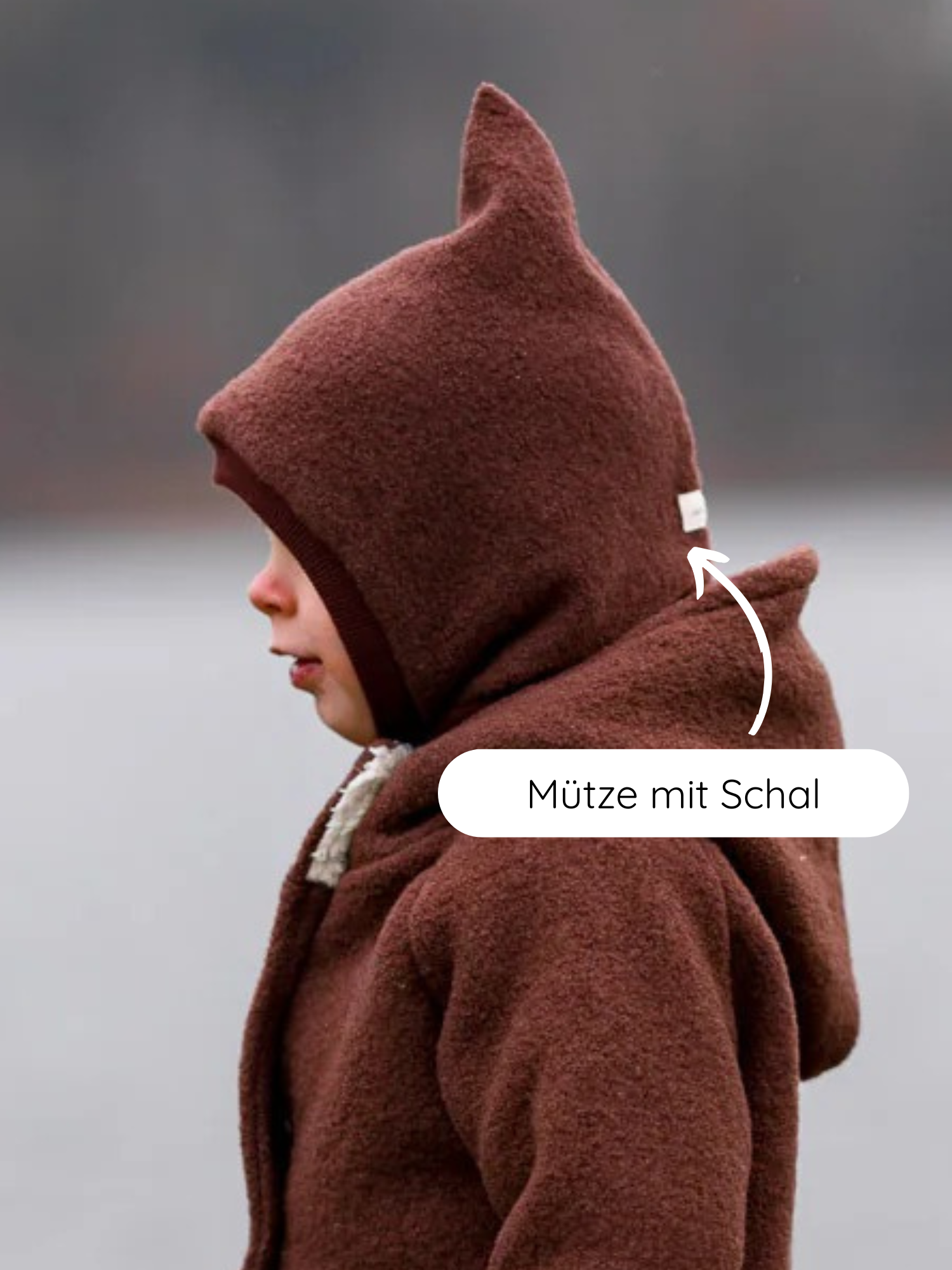 Kleinkind im Profil trägt Schlupfmütze Wollwalk - Schoko mit spitzer Kapuze und integriertem Schal, perfekt sitzend und warm. Mütze mit Schal steht im Bild.