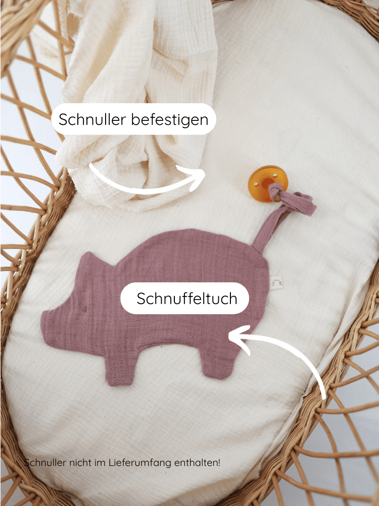 Schmusetuch/Schnullerband Schweinchen aus Baumwoll-Musselin in einem Moses-Korb, begleitet von einer cremefarbenen Muslin-Decke. Pig-förmiges Schnuffeltuch mit Schnuller-Schlaufe, Hinweis: Schnuller nicht enthalten.