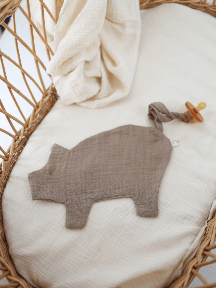 Schmusetuch/Schnullerband Schweinchen Baumwoll-Musselin - Praline: Ein flaches, schweinförmiges Komforttuch aus braun-grauem Leinen mit geknotetem Schwanz, der einen Schnuller hält, auf weichem, weißem Muslin in einem Korb liegend.