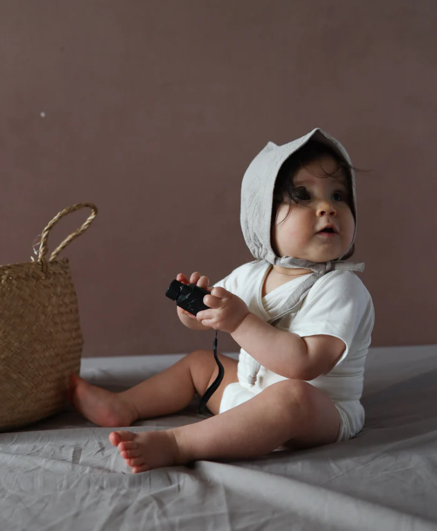 Ein Baby trägt einen Kurzarm Loopbody Bambusviskose, sitzt auf einer weichen grauen Decke und hält neugierig eine kleine Kamera. Neben ihm steht ein geflochtener Strohkörbchen.