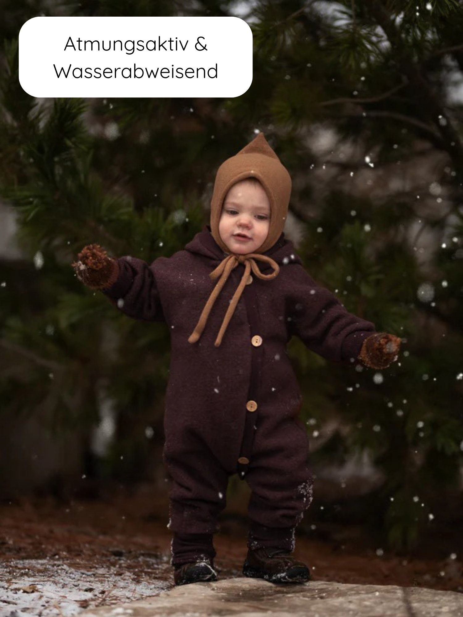 Kleinkind im Winter Wollwalk Overall mit Teddyfutter - Malve, trägt braunen Einteiler mit Holzknöpfen, passenden Fäustlingen und Stiefeln, steht im leichten Schneefall, umgeben von immergrünen Bäumen.