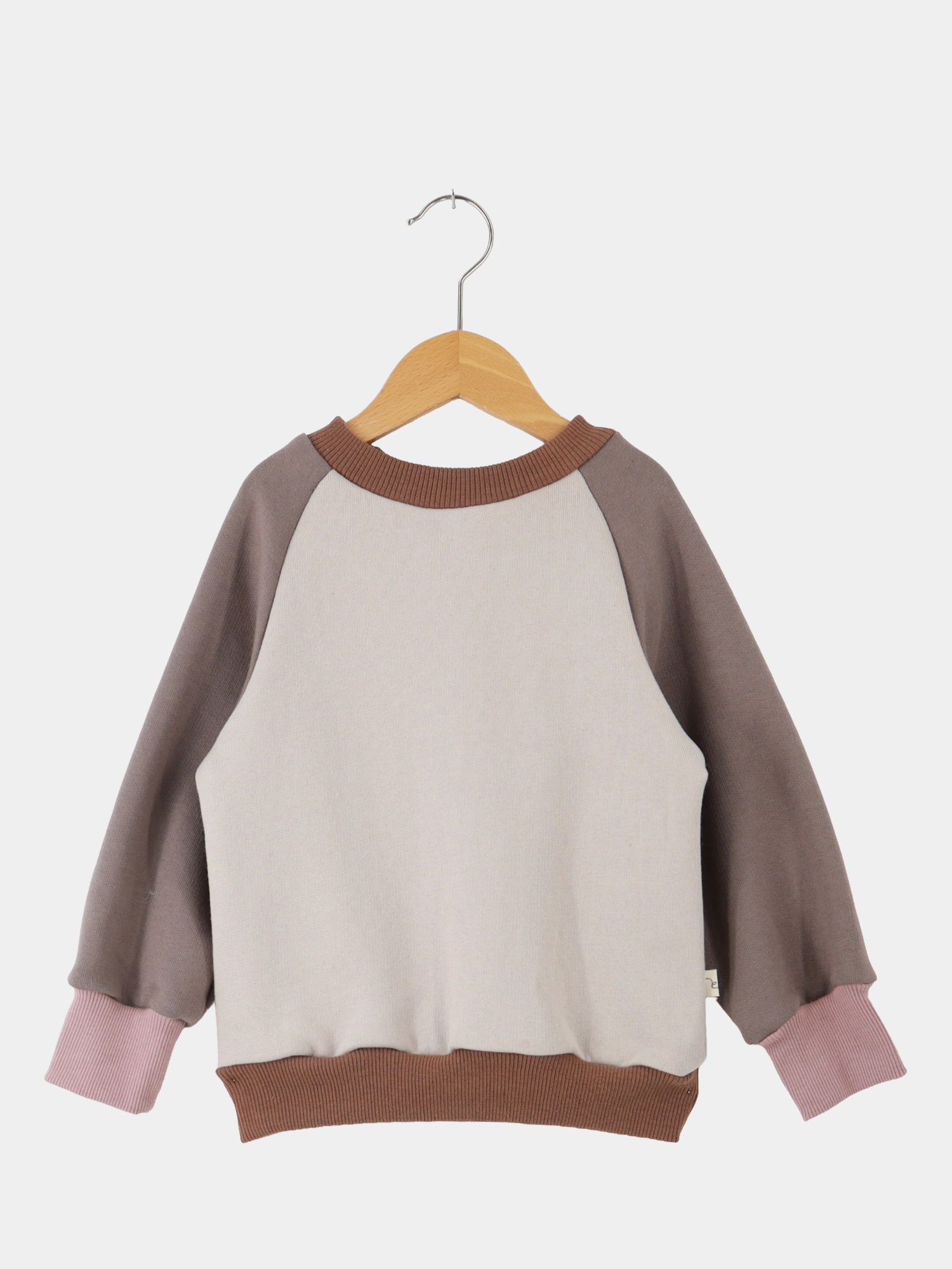 Upcycling-Pulli Bio-Sweat - SanfteErde: Kinderpullover mit langen Ärmeln, beige Körper, braune Ärmel, kontrastierende rosa Bündchen, aus GOTS-zertifizierter Bio-Baumwolle, aus Stoffresten gefertigt, auf Holzbügel vor weißem Hintergrund.