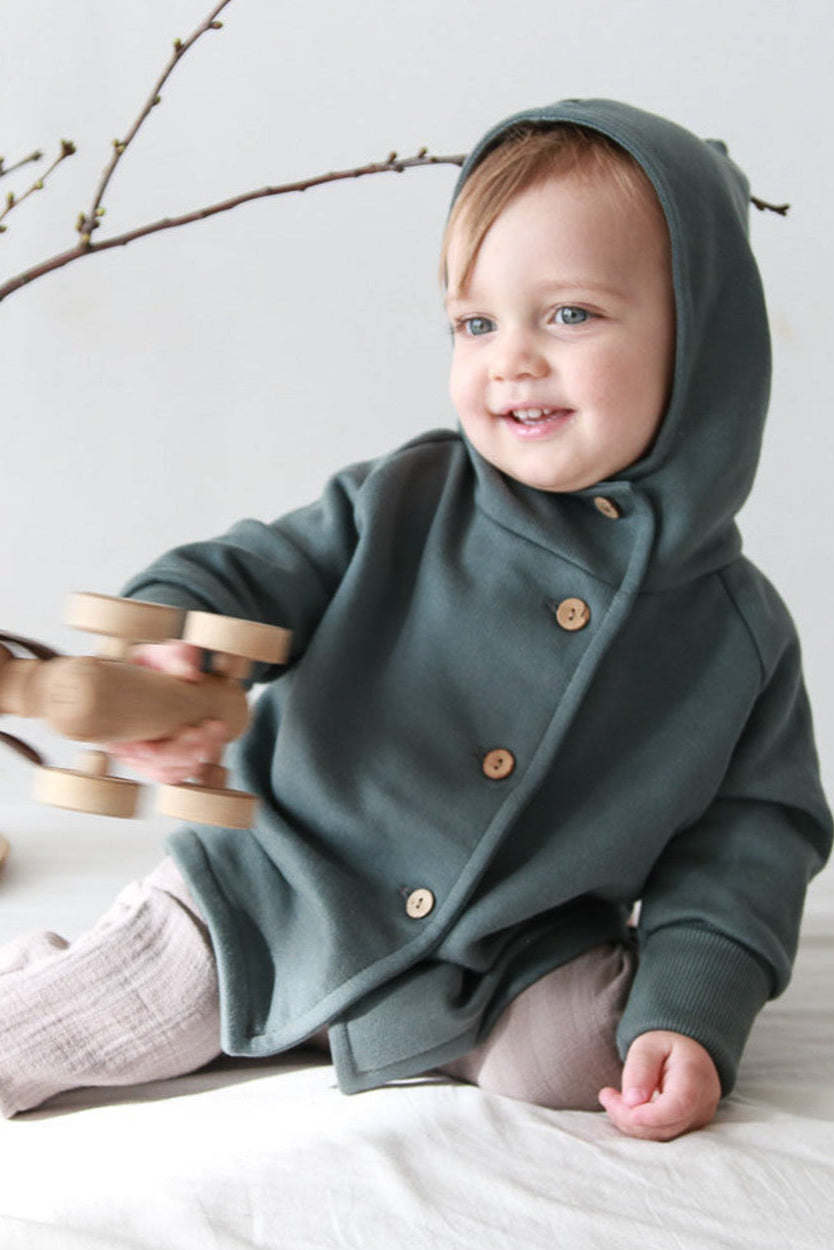 Ein Baby sitzt auf dem Boden und hält ein Holzspielzeug, während es eine Wichteljacke Bio-Sweat - Taube von PETIT COCHON Berlin trägt.
