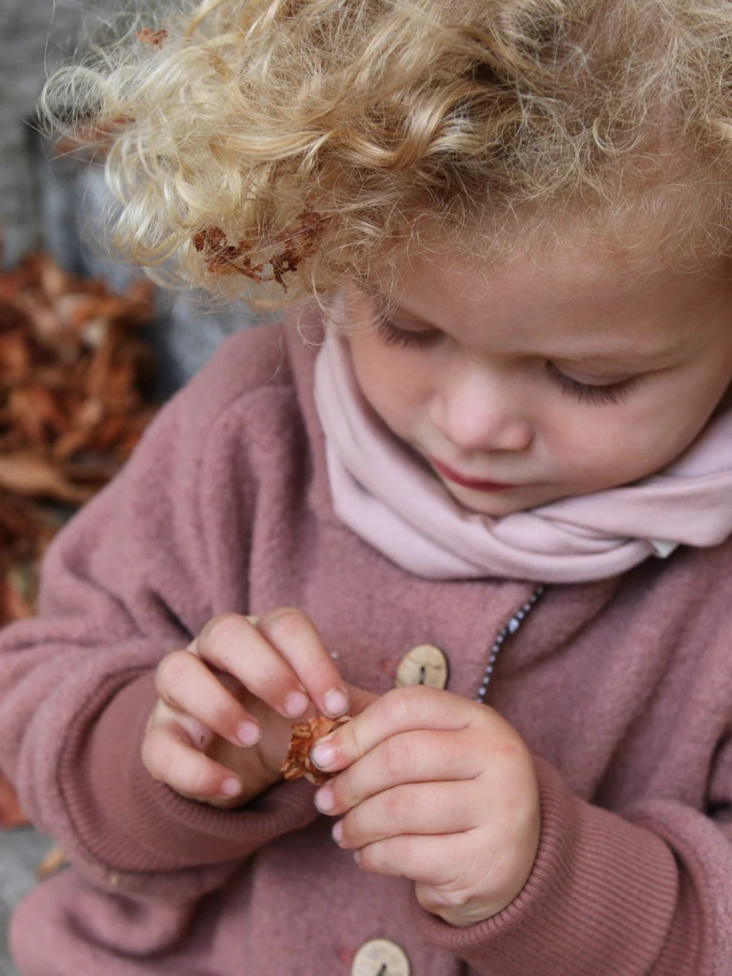 Ein Kind trägt die Wichteljacke Wollwalk - Zimt, erkundet neugierig Herbstlaub. Die Jacke bietet Bewegungsfreiheit und ist aus hochwertigem Wollwalk mit Bio-Jersey-Futter, ideal für kalte Jahreszeiten.