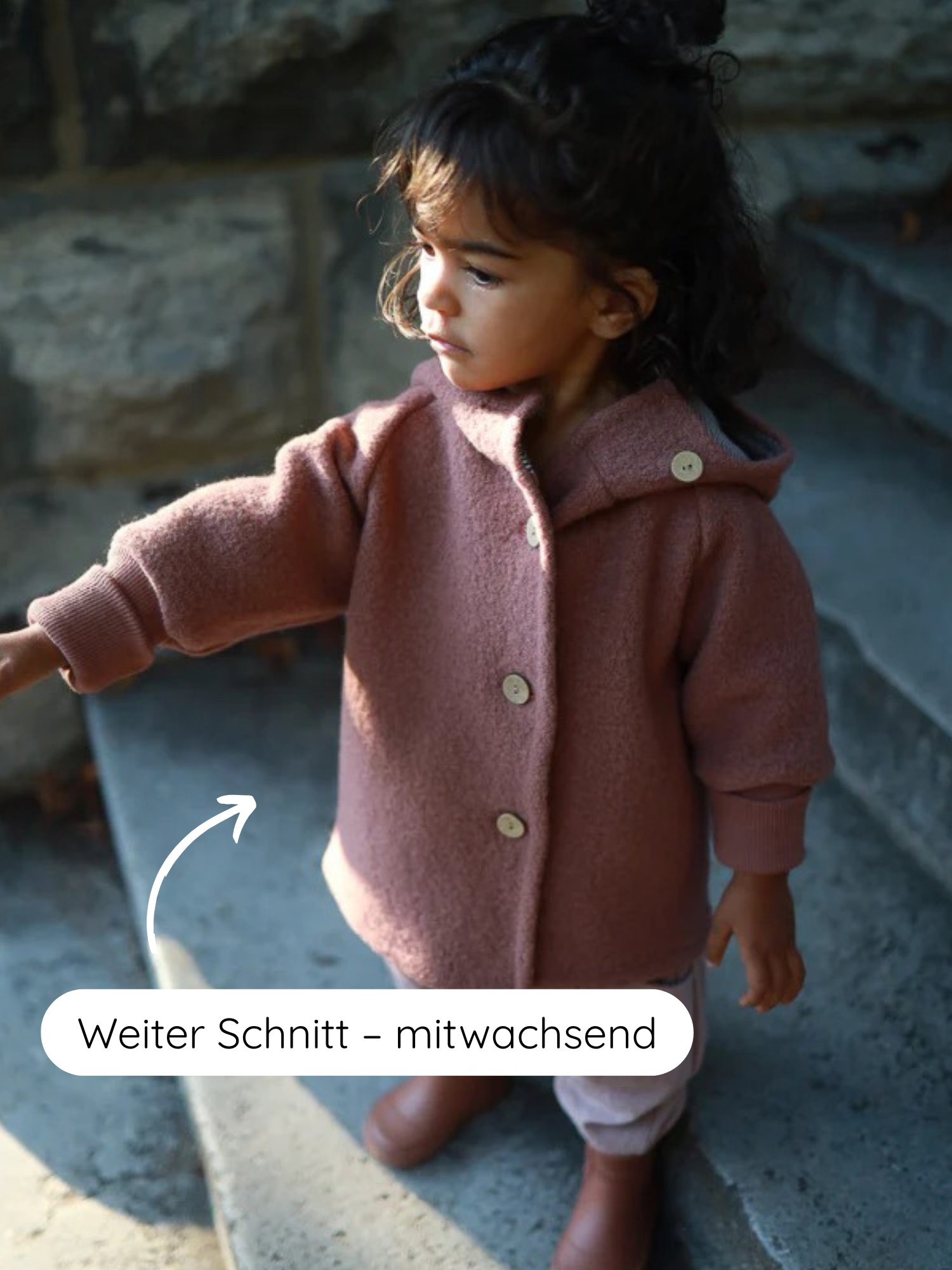 Ein Kleinkind trägt die Wichteljacke Wollwalk - Zimt mit Kapuze, die großzügig geschnitten ist. Der Pfeil zeigt den mitwachsenden Schnitt, ideal für Herbst bis Winter.
