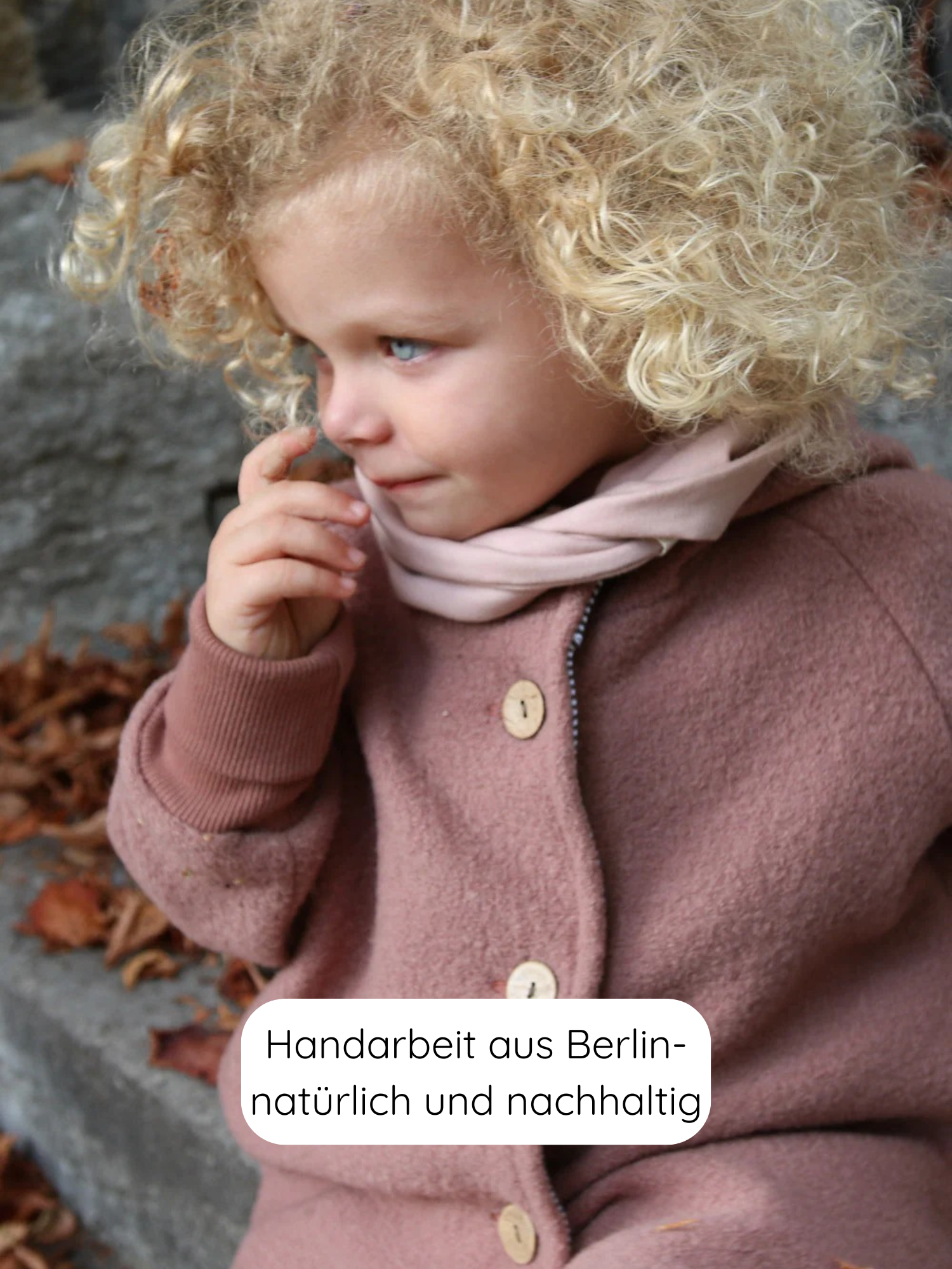 Ein Kind sitzt draußen auf einer Steintreppe mit Herbstblättern, trägt die Wichteljacke Wollwalk - Zimt und einen rosa Schal. Die Jacke ist handgefertigt, natürlich und nachhaltig aus Berlin.