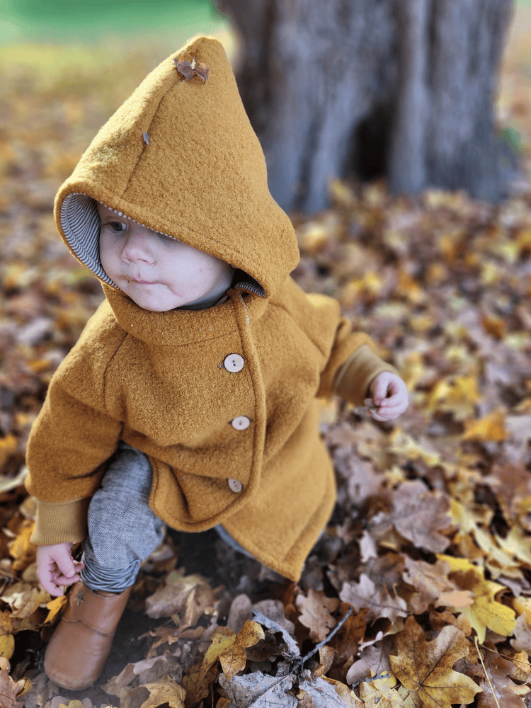 Ein Kleinkind trägt eine Wichteljacke Wollwalk - Honig mit Holzknöpfen, während es in Herbstlaub hockt. Die Jacke hat eine Kapuze und krempelbare Bündchen, bietet Wärme und Bewegungsfreiheit.