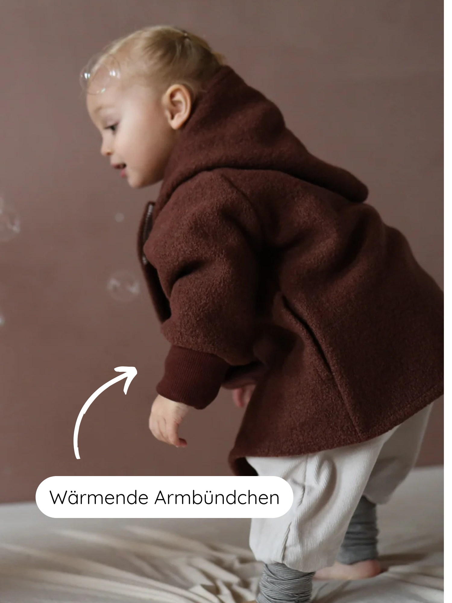 Kleinkind trägt Wichteljacke Wollwalk - Schoko mit Bio-Jersey-Futter. Spielt mit Seifenblasen, weiße Pfeil zeigt auf Wärmende Armbündchen an den gerippten Bündchen. Ideal für Herbst, Winter und Frühling.