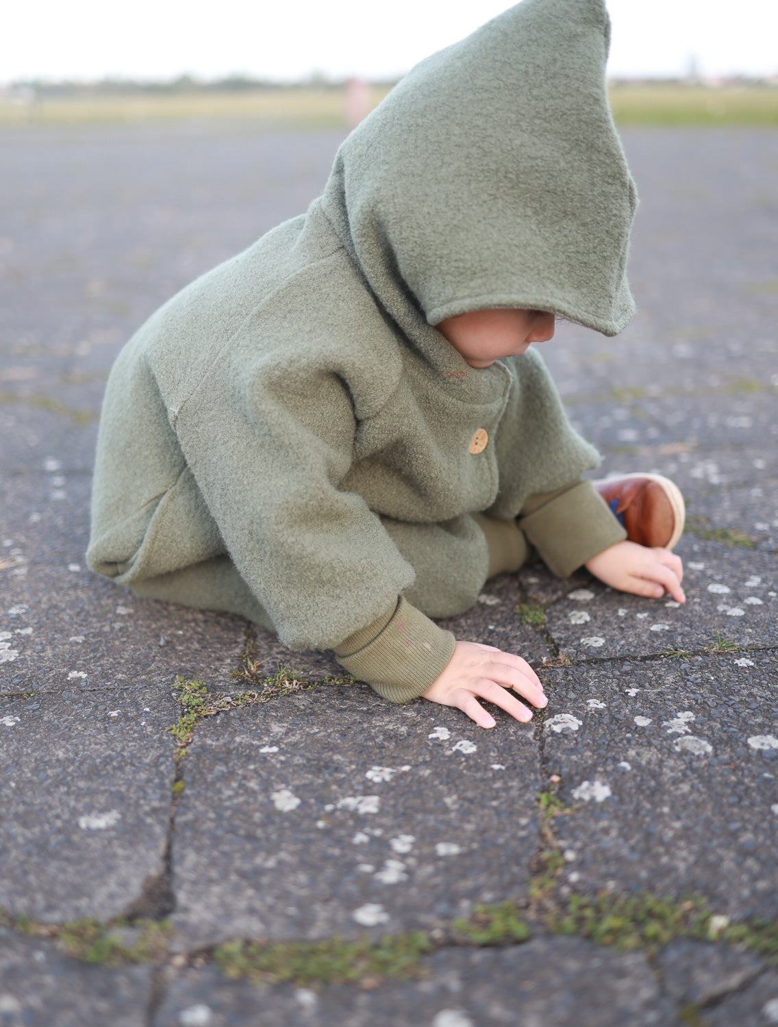 Ein Baby trägt die Wichteljacke Wollwalk - Pistazie auf einem Steinboden; die Jacke bietet Bewegungsfreiheit und temperaturregulierende Eigenschaften, gefüttert mit Bio-Baumwoll-Jersey für Komfort. nachhaltige Kinderjacke aus Wollwalk