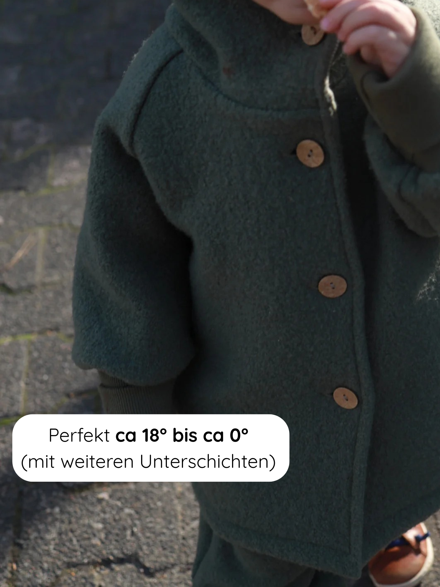 Kind mit Wollwalk-Wichteljacke - Pistazie, trägt eine grüne Jacke mit Holzknöpfen, hält Snack. Perfekt für 18°C bis 0°C mit Unterschichten. Hochwertig, gefüttert, mitwachsend, ideal für Herbst bis Frühling.