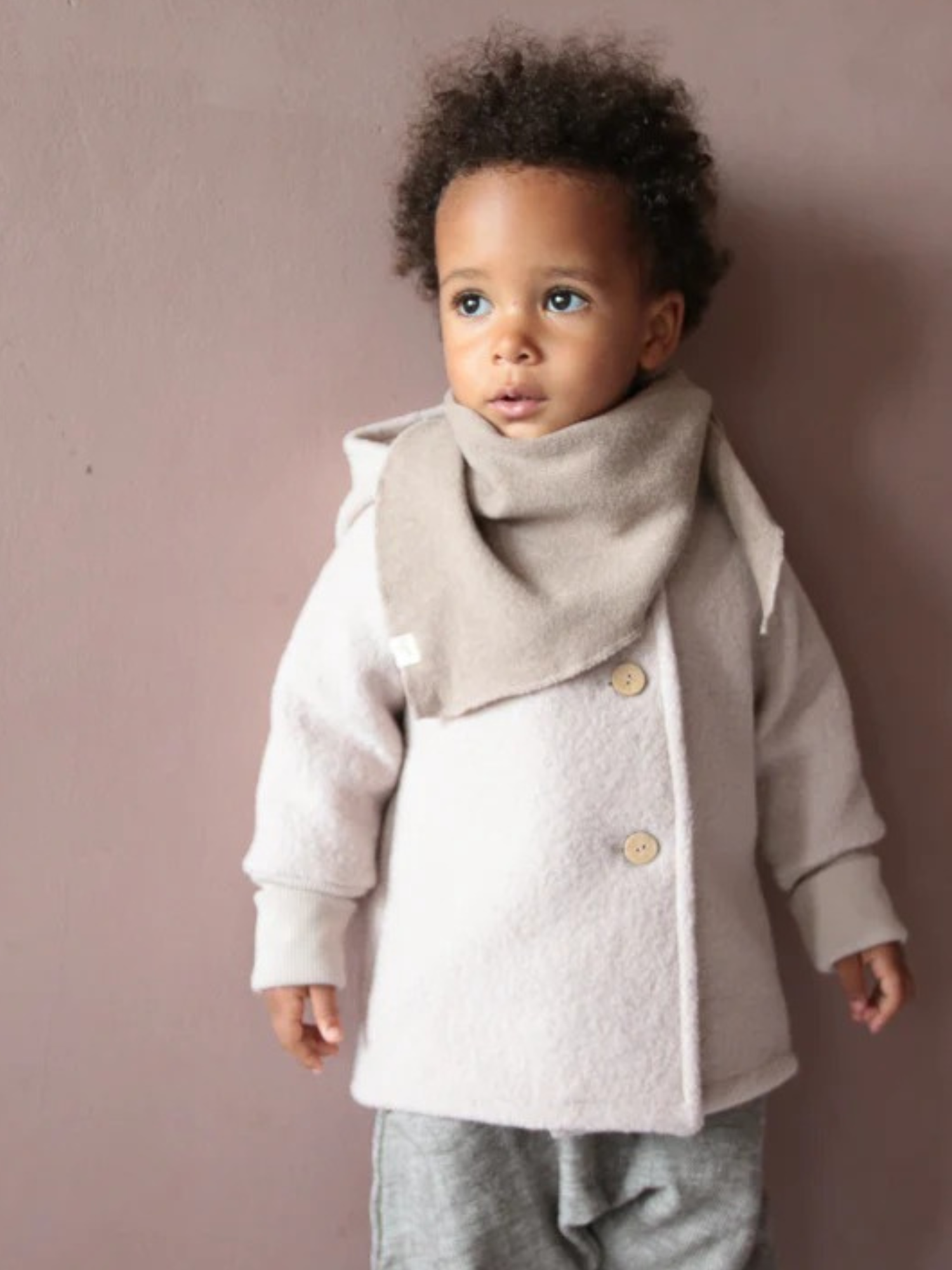 Ein Kleinkind trägt die Wichteljacke Wollwalk - Sand, eine beige Knopfjacke mit passendem Schal, über grauen Hosen. Die Jacke bietet Bewegungsfreiheit und ist ideal für kühlere Jahreszeiten.