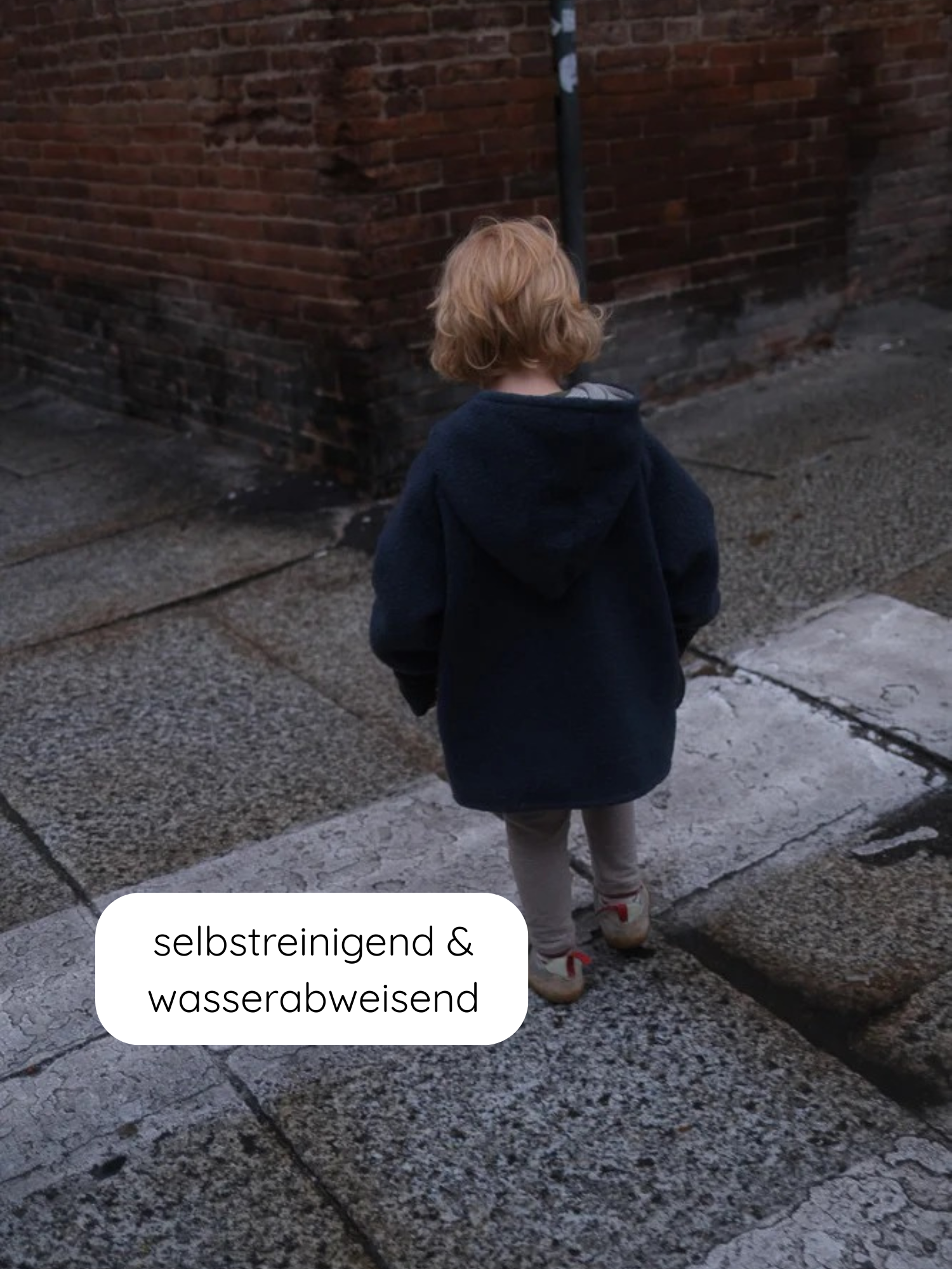 Kleinkind von hinten in einer übergroßen Wichteljacke Wollwalk - Marine, mit Kapuze auf einem Pflasterweg vor einer Backsteinmauer stehend, mit der Aufschrift selbstreinigend & wasserabweisend.