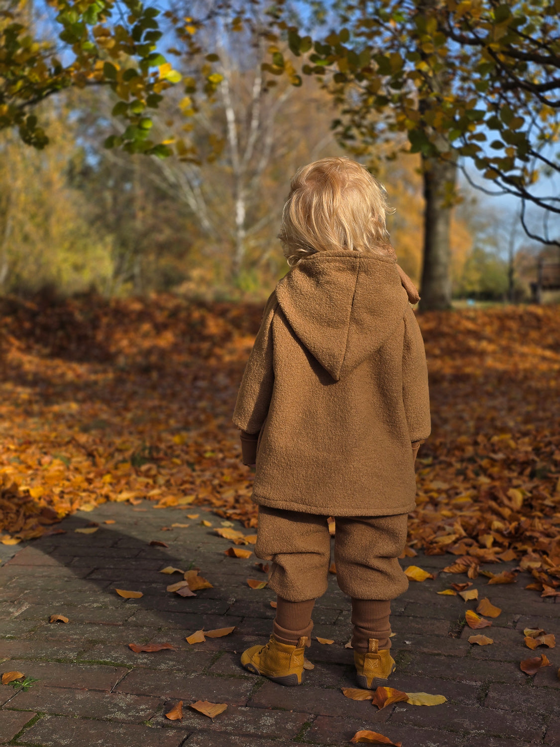 Kind in der Wichteljacke Wollwalk - Kamel steht auf einem Laubweg. Die Jacke bietet große Bewegungsfreiheit und ist für Herbst bis Winter ideal. Sie besteht aus hochwertigem, gefüttertem Wollwalk. nachhaltige Wollwalk Jacke für Kinder