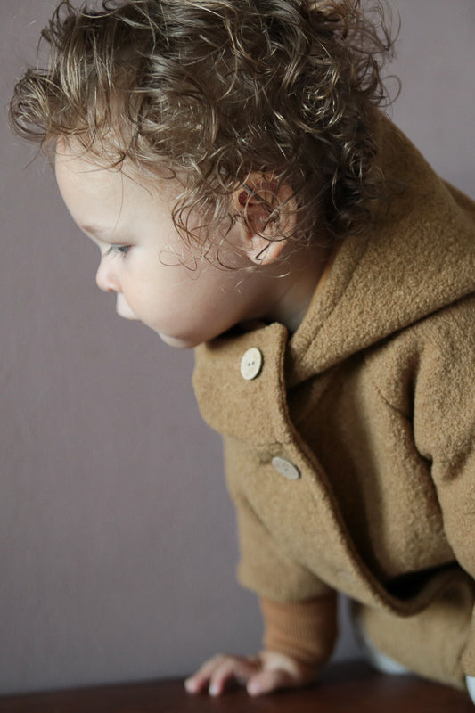 Kleinkind trägt die Wichteljacke Wollwalk - Kamel, hergestellt aus hochwertigem Wollwalk mit Bio-Jersey-Futter, ideal für Herbst und Winter, bietet Bewegungsfreiheit und temperaturregulierende Eigenschaften.